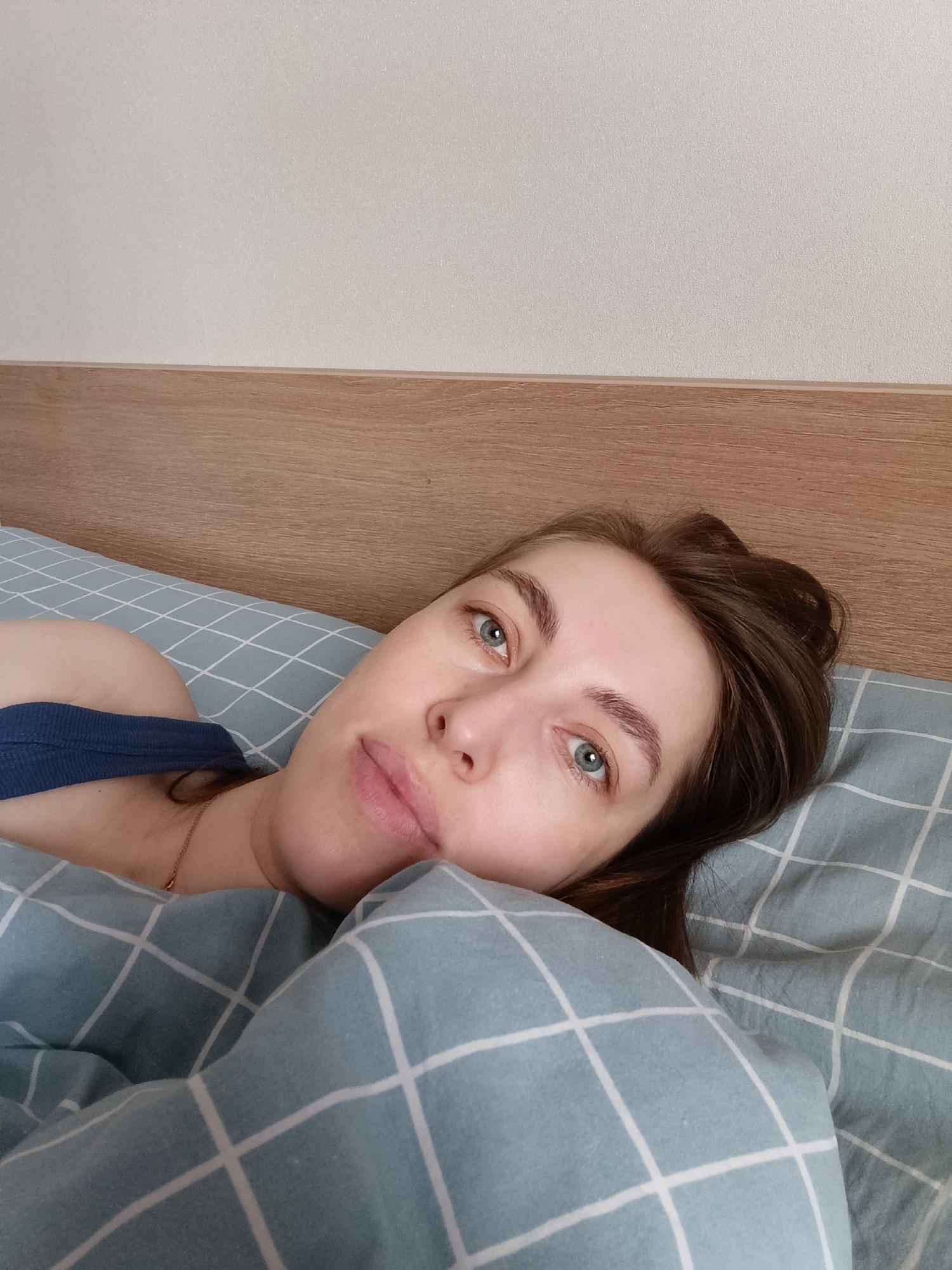 A young woman is pictured lying in bed, looking directly at the camera with a calm, neutral expression. She has light skin, blue-green eyes, and brown hair. Her hair is casually spread around her head as she lies on her side. She is wearing a blue tank top, with a delicate gold chain visible around her neck.
She is partially covered by a light blue duvet or blanket, which features a distinct white grid pattern. A pillow with the same pattern is beneath her head. Behind her, a light wooden headboard runs horizontally, separating the bed from a plain, light-colored wall that occupies the upper portion of the frame. The indoor setting suggests a bedroom.
The lighting is natural and soft, indicative of daytime, possibly morning, creating a relaxed and intimate atmosphere. No specific activity is taking place beyond her resting or posing in bed. There is no visible text in the image. The scene is situated in Chaikovsky, Russia.