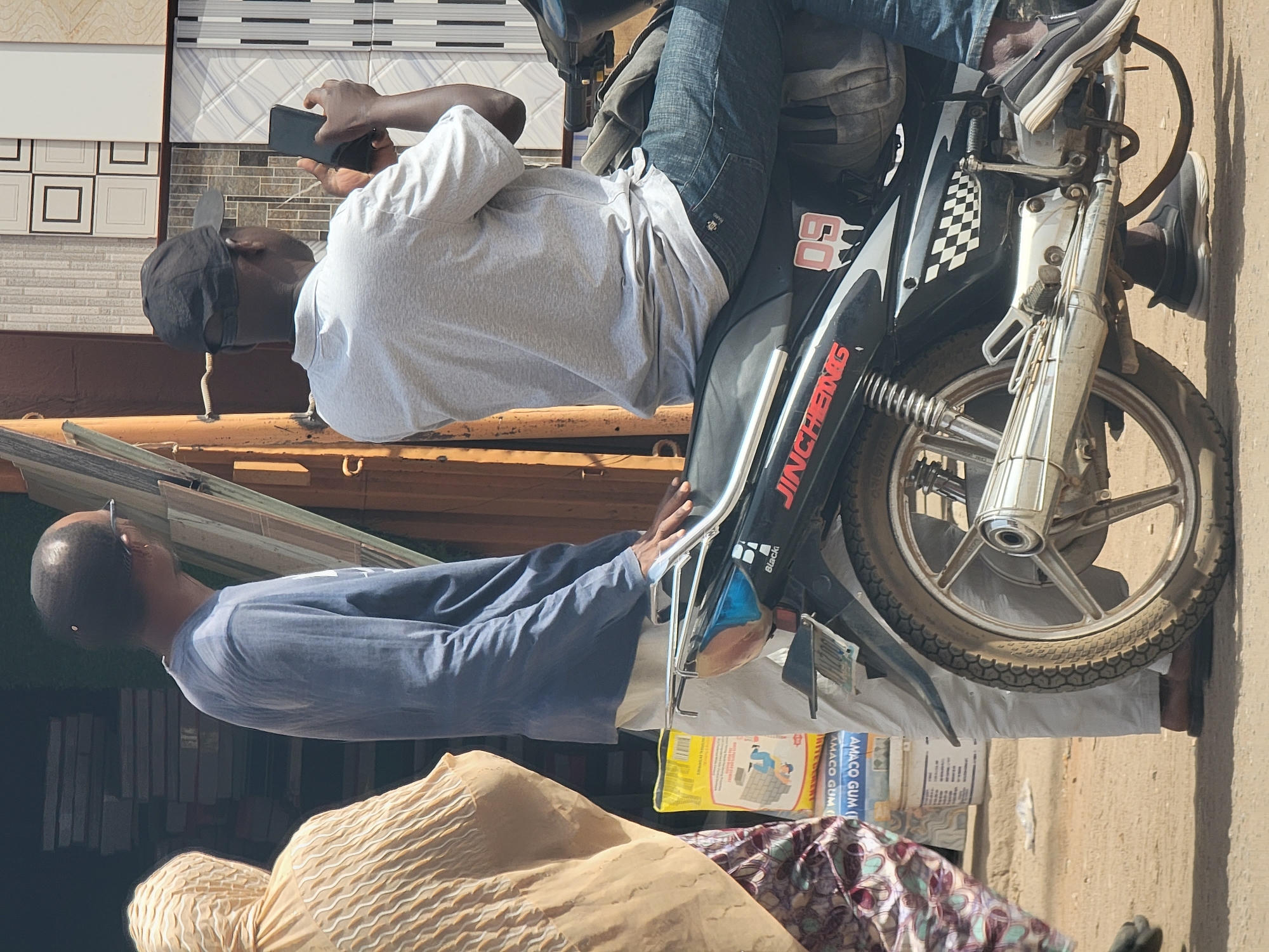 A man wearing a gray shirt, blue jeans, and sneakers sits on a black motorcycle on a city street in Maiburiji, Nigeria. He holds a phone in his hand. Another man wearing a blue shirt, white pants, and a brown robe stands next to the motorcycle. There are boxes of Amaaco Gum and other products on the ground next to the motorcycle. The street is paved. In the background is a building with rows of tiles displayed on the wall.