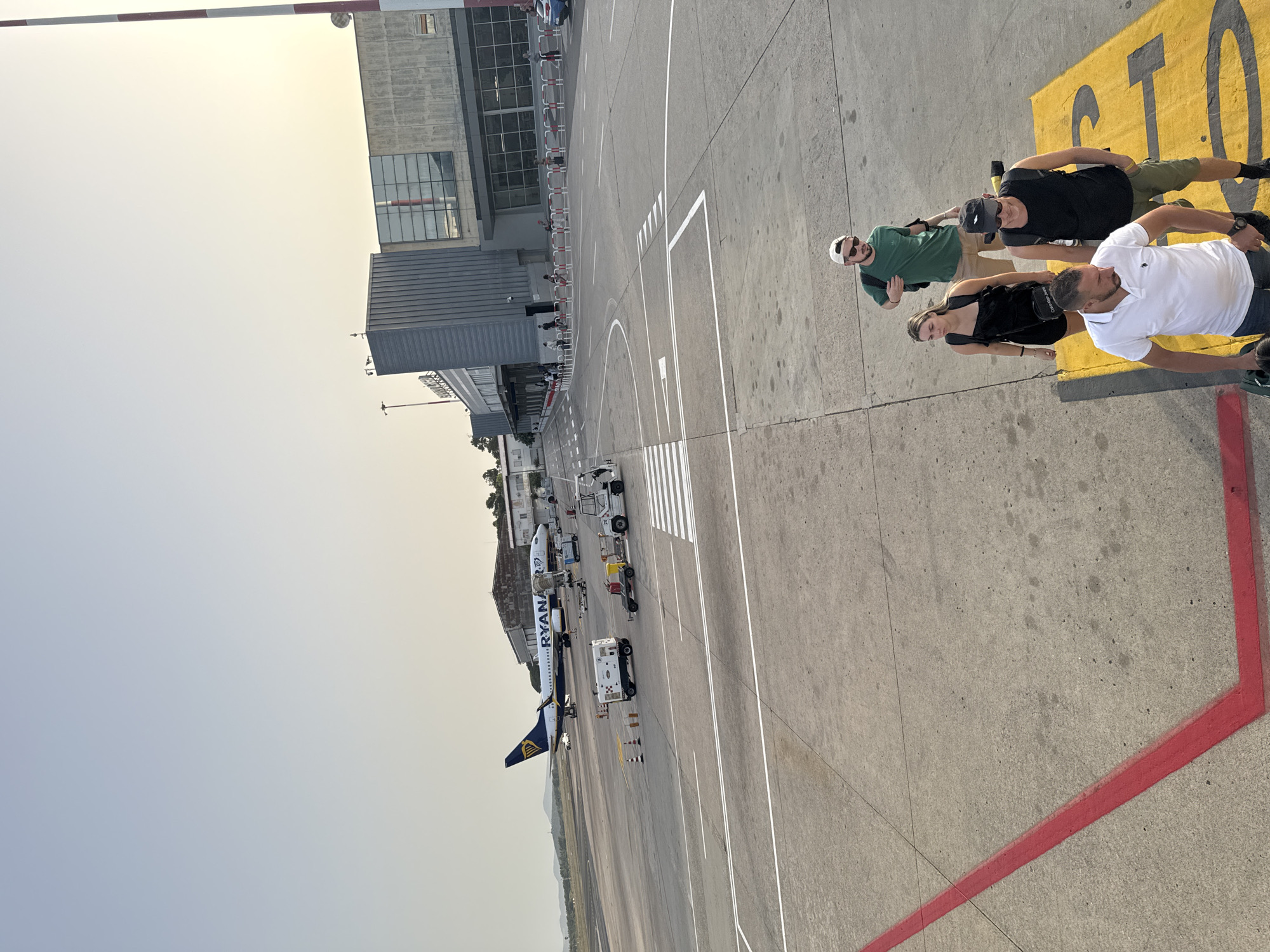 The image depicts an airport tarmac scene in Alghero, Italy. A Ryanair airplane is parked on the tarmac, with ground support equipment nearby. Several individuals are walking on a jet bridge, appearing to board or disembark the aircraft. The airport terminal building is visible in the background, characterized by a modern architectural style with large windows. The sky is clear, suggesting fair weather conditions. The scene captures a typical moment of air travel activity at an airport.