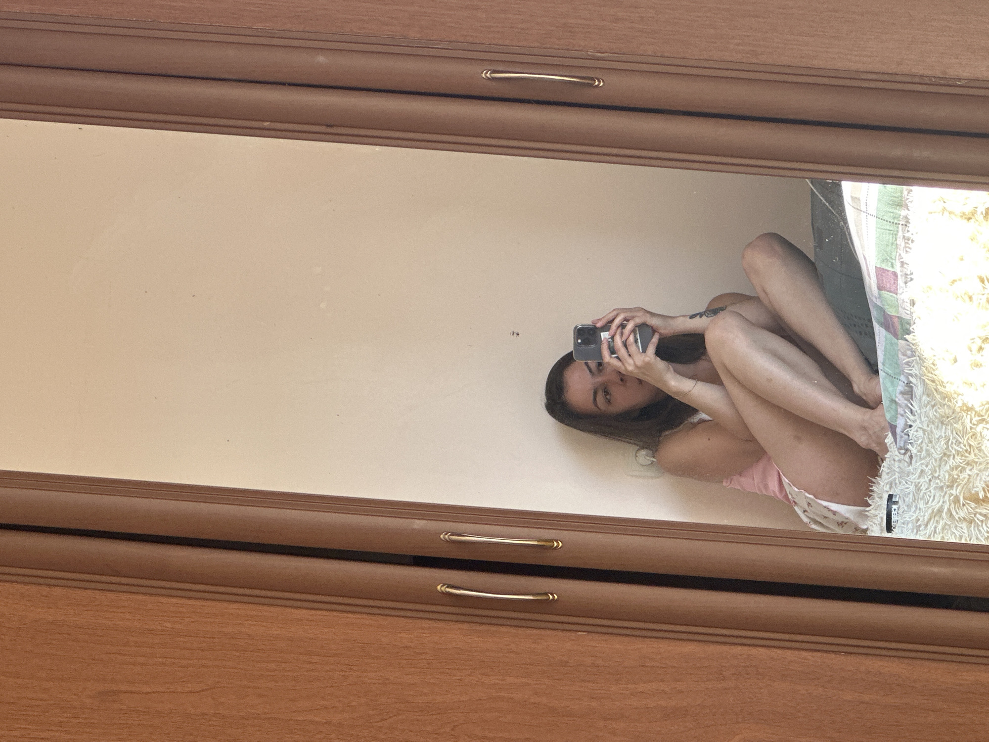 A young woman with dark hair is sitting on a bed, reflected in a mirror. She is wearing a pink tank top and patterned shorts. She is holding a smartphone in her hands, taking a picture of herself. The background is a plain wall. The mirror is framed by a wooden wardrobe. The image was taken in Gyumri, Armenia.
