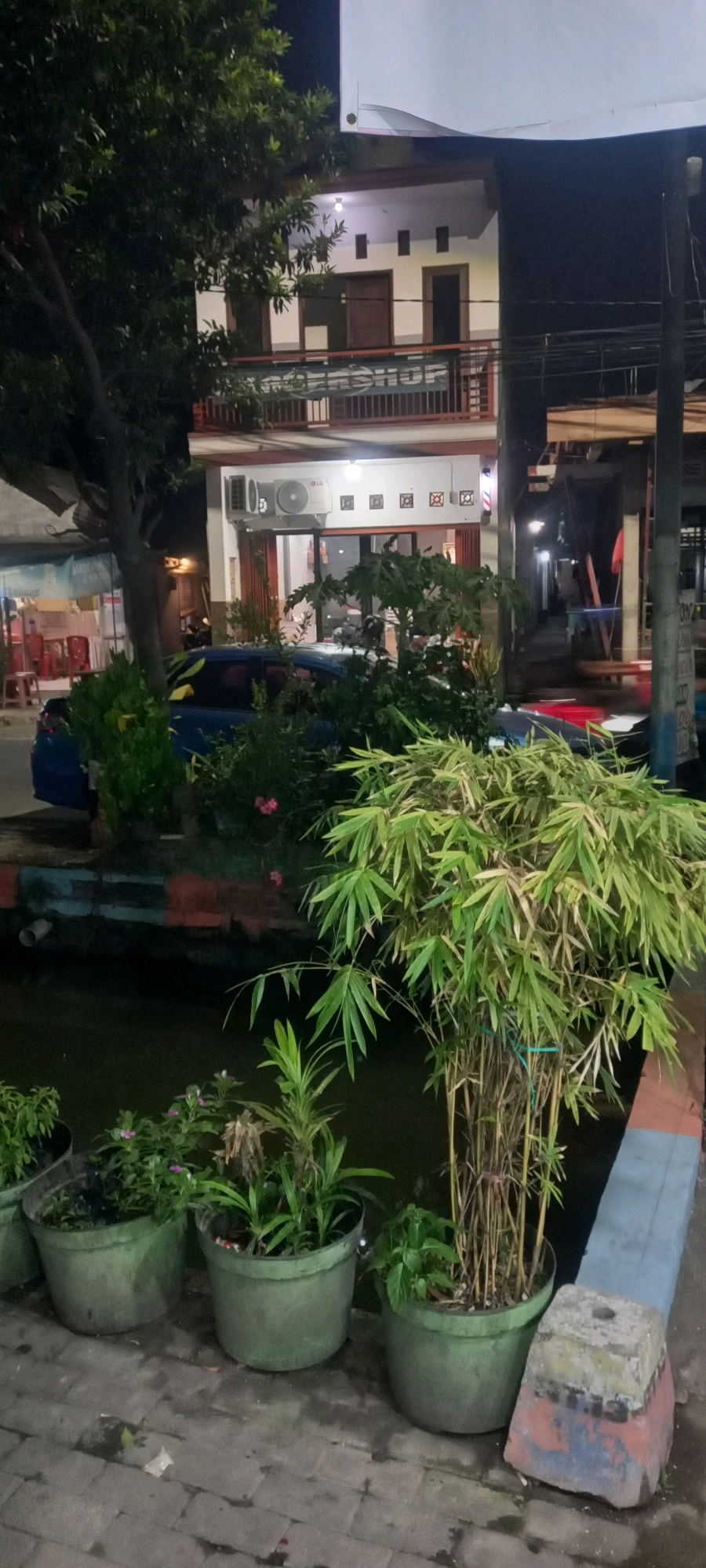 In Sedati Agung, Indonesia, at night, a street scene unfolds with a two-story building dominating the background. The building features white walls, dark trim around windows and doors, and balconies on both levels. An air conditioning unit is visible on the lower floor, along with several decorative square frames. A barber pole sign is mounted near the right side of the building's entrance. A blue car is parked in front of the building, partially obscured by greenery.
In the foreground, several potted plants are arranged along a walkway paved with stone. A large bamboo plant in a pot stands prominently on the right. Other smaller plants are in similar pots. A narrow channel of dark water runs alongside the walkway, bordered by a painted concrete edge with alternating bands of color. A tree with dark foliage is positioned to the left, casting shadows. The overall lighting suggests artificial light sources are active.