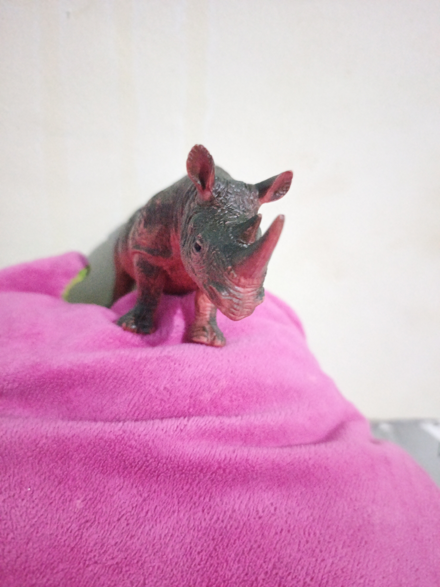 A toy rhinoceros figurine is positioned on a plush, bright pink object, possibly a pillow or blanket, in a close-up, high-angle shot. The rhinoceros is dark gray with reddish-brown highlights and has a prominent horn. The background is a plain, off-white wall. The lighting is soft, casting gentle shadows. There is no identifiable location within South Jakarta, Indonesia.