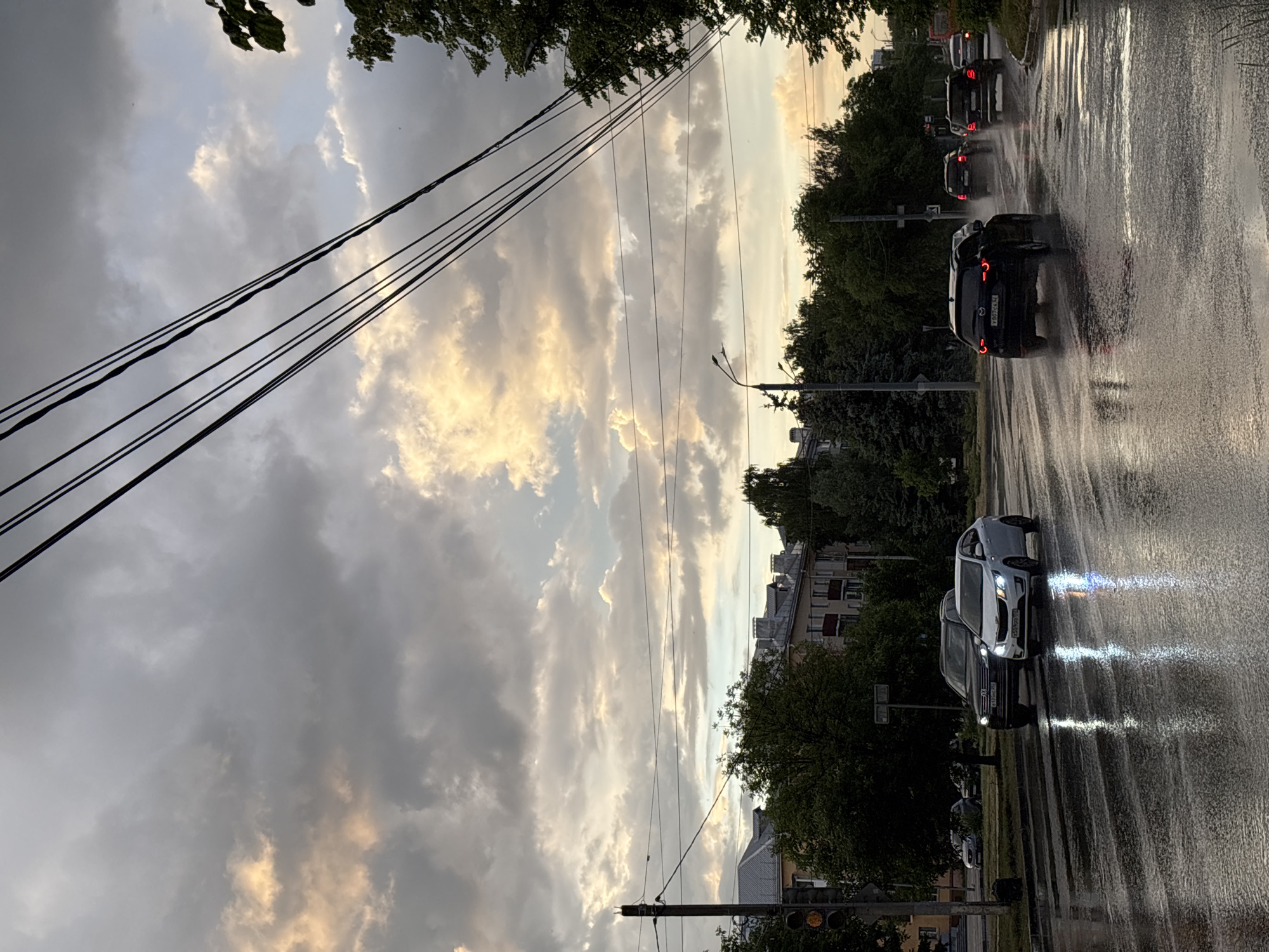 The image captures a street scene in Nizhny Novgorod, Russia, under a dramatic sky filled with a mix of dark and light-colored clouds. The sun's rays are breaking through, creating illuminated patches in the sky. The street is wet and reflects the lights of the vehicles driving on it, suggesting recent rainfall or ongoing drizzle.
Several cars are visible on the road. In the foreground, a dark-colored SUV and a white sedan are driving. Further down the street, more cars are seen, also moving through the wet conditions. Trees line the sides of the street, and buildings with distinctive chimneys are visible in the background, characteristic of urban architecture. Overhead, power lines stretch across the frame, adding to the urban feel of the scene. Traffic lights are also present, indicating a populated and active area. The overall atmosphere is one of a city road during a period of inclement weather, with the dramatic sky creating a striking visual backdrop.