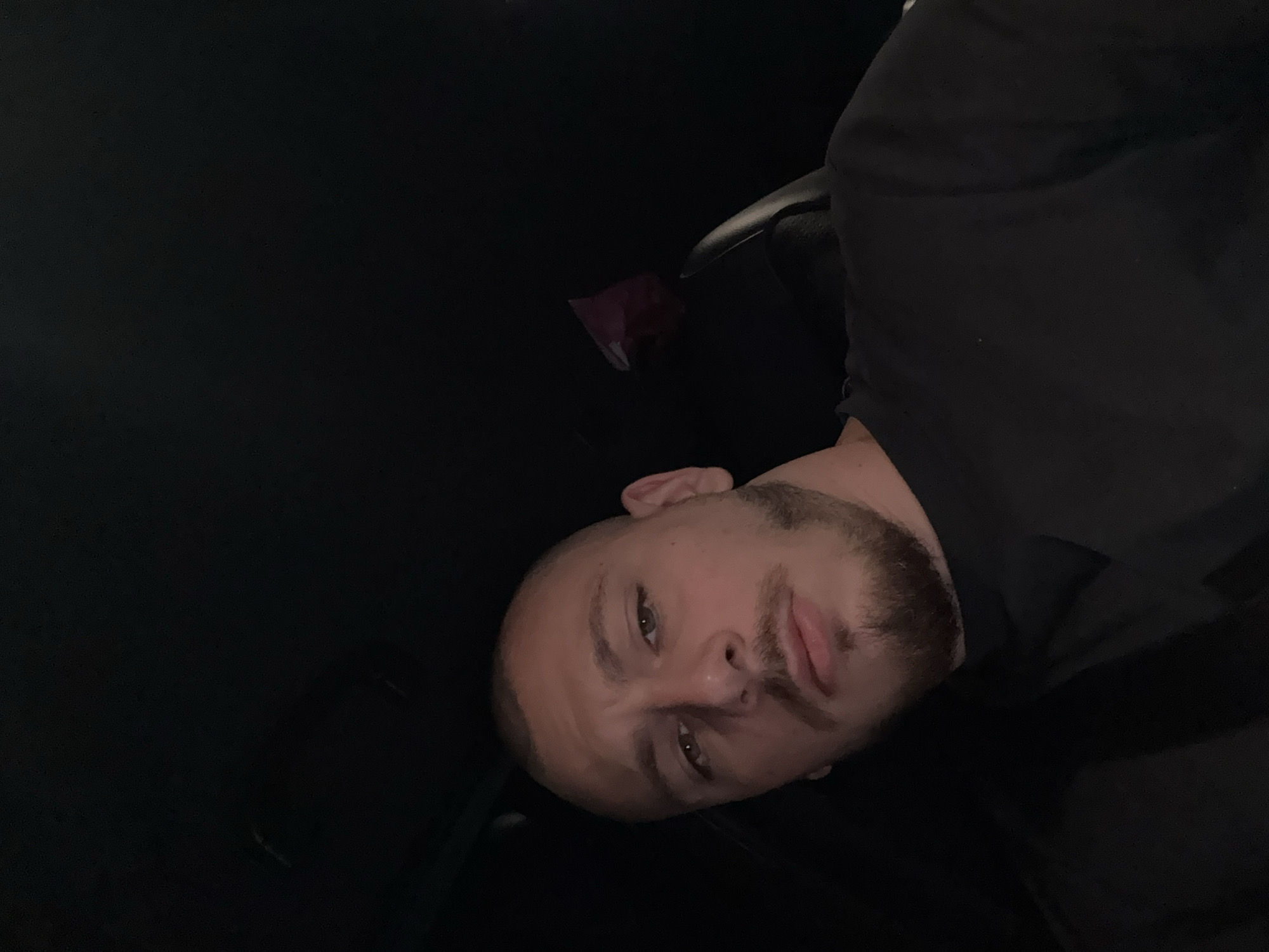 The image is a close-up, low-angle selfie of a man with a shaved head and a goatee, wearing a black t-shirt. He is looking directly at the camera with a neutral expression. The background is dark and appears to be the interior of a vehicle, with a hint of a purple object and part of a headrest visible. The lighting is dim, with the man's face illuminated from the front. The setting is not discernible as being in Stara Zagora, Bulgaria.