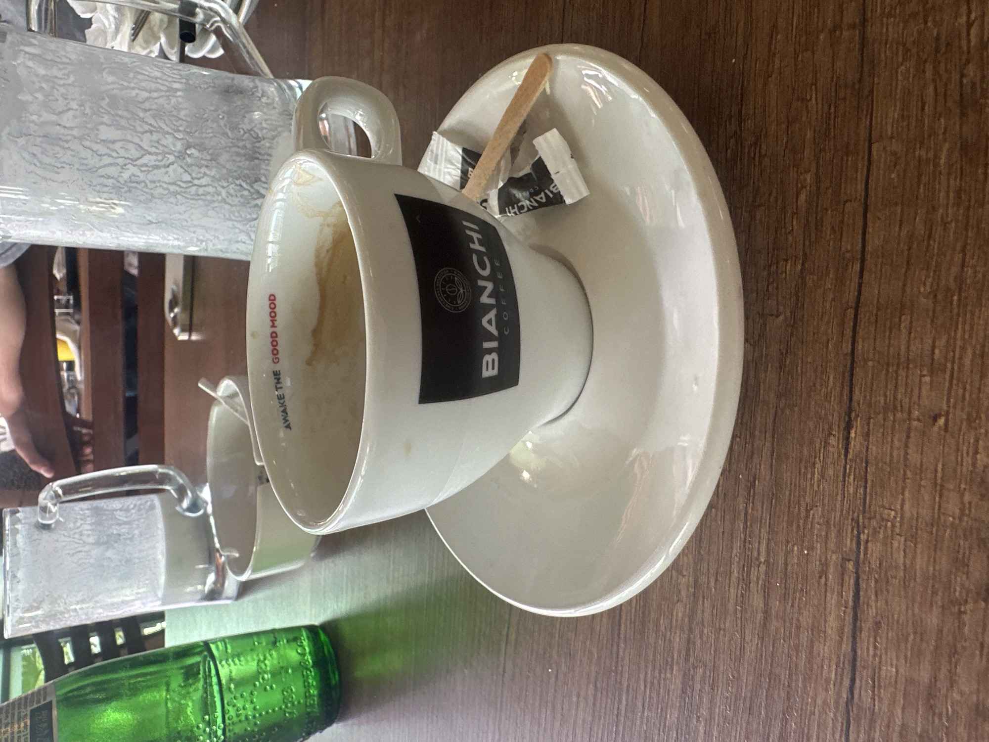 The image shows a coffee cup, saucer, sugar packets, and a wooden stirring stick on a wooden table. The cup is branded "Bianchi Coffee" and has the phrase "Awake the Good Mood" written on it. The location is Ivaylovgrad, Bulgaria, inferred from the "Bianchi Coffee" branding, which has a local presence. A green bottle and another glass container are also visible. There are no people explicitly visible in the primary focus, but a hand is visible in the background.