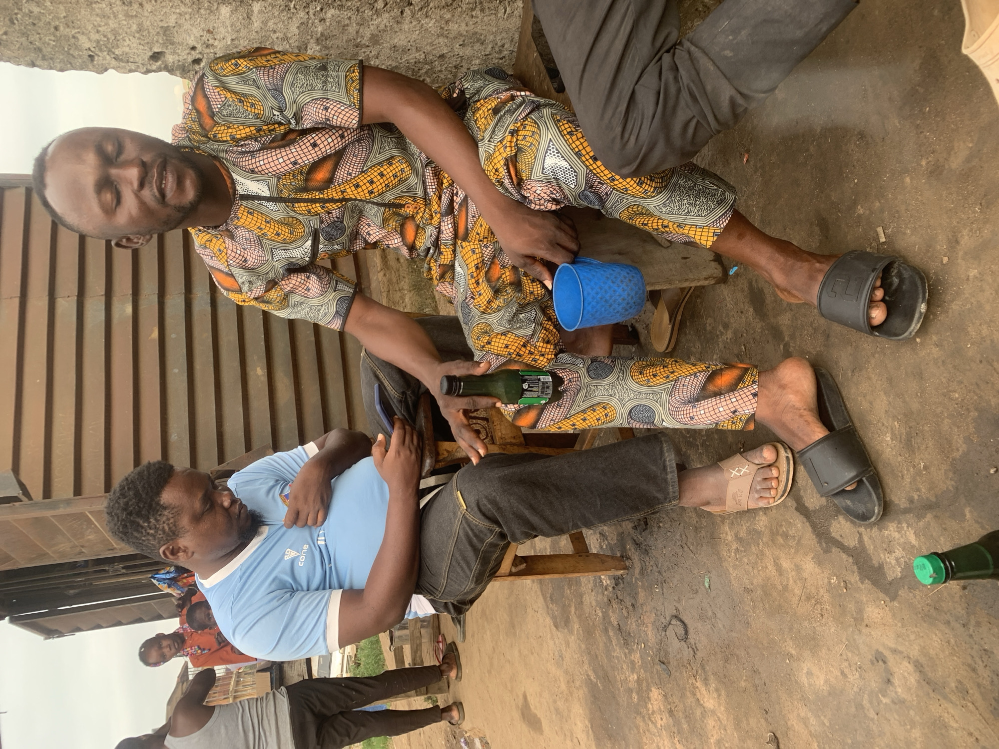 The image depicts an outdoor scene in Shagamu, Nigeria. Two men are seated, facing away from the viewer and towards each other. The man on the left, wearing a light blue t-shirt and dark jeans, is looking down at his phone, which he holds in his right hand. He is seated on a wooden stool. The man on the right, wearing a brightly patterned dashiki and dark pants, has his eyes closed and appears to be resting. He is holding a small blue cup in his left hand. A bottle of green liquid and a partial bottle with a green cap are visible on the ground in the foreground. The environment appears to be a dusty, unpaved area, possibly a yard or a street, with a corrugated metal structure in the background and a rough concrete wall to the right. The overall impression is that of a casual gathering or moment of repose.