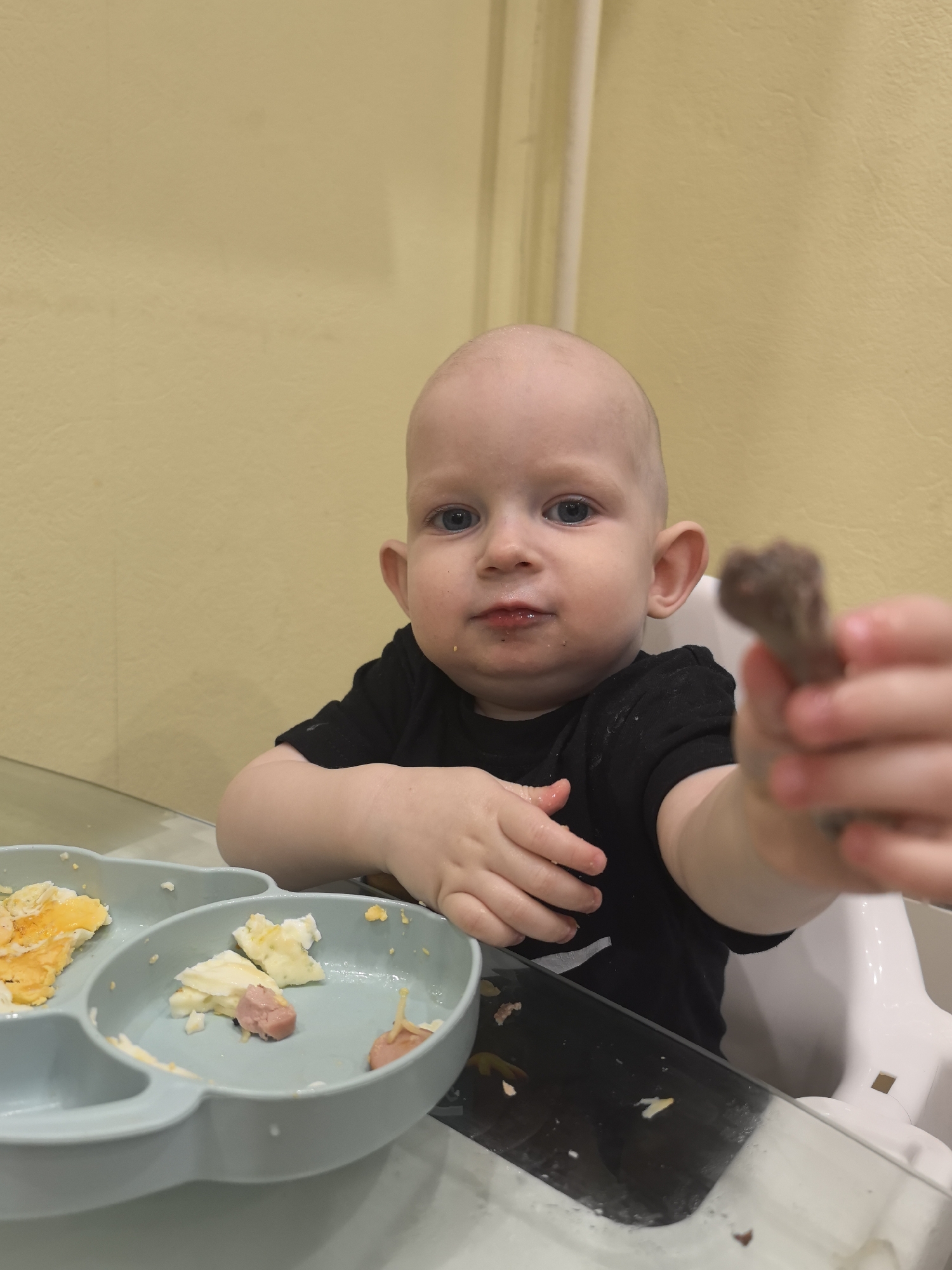 A baby with a bald head, wearing a black T-shirt, sits at a highchair facing the camera, holding a piece of food in their right hand. A gray divided plate containing scrambled eggs and sliced sausage sits to their left. The environment appears to be a domestic setting with yellow walls. The location cannot be definitively determined to be Moscow, Russia, without additional information.