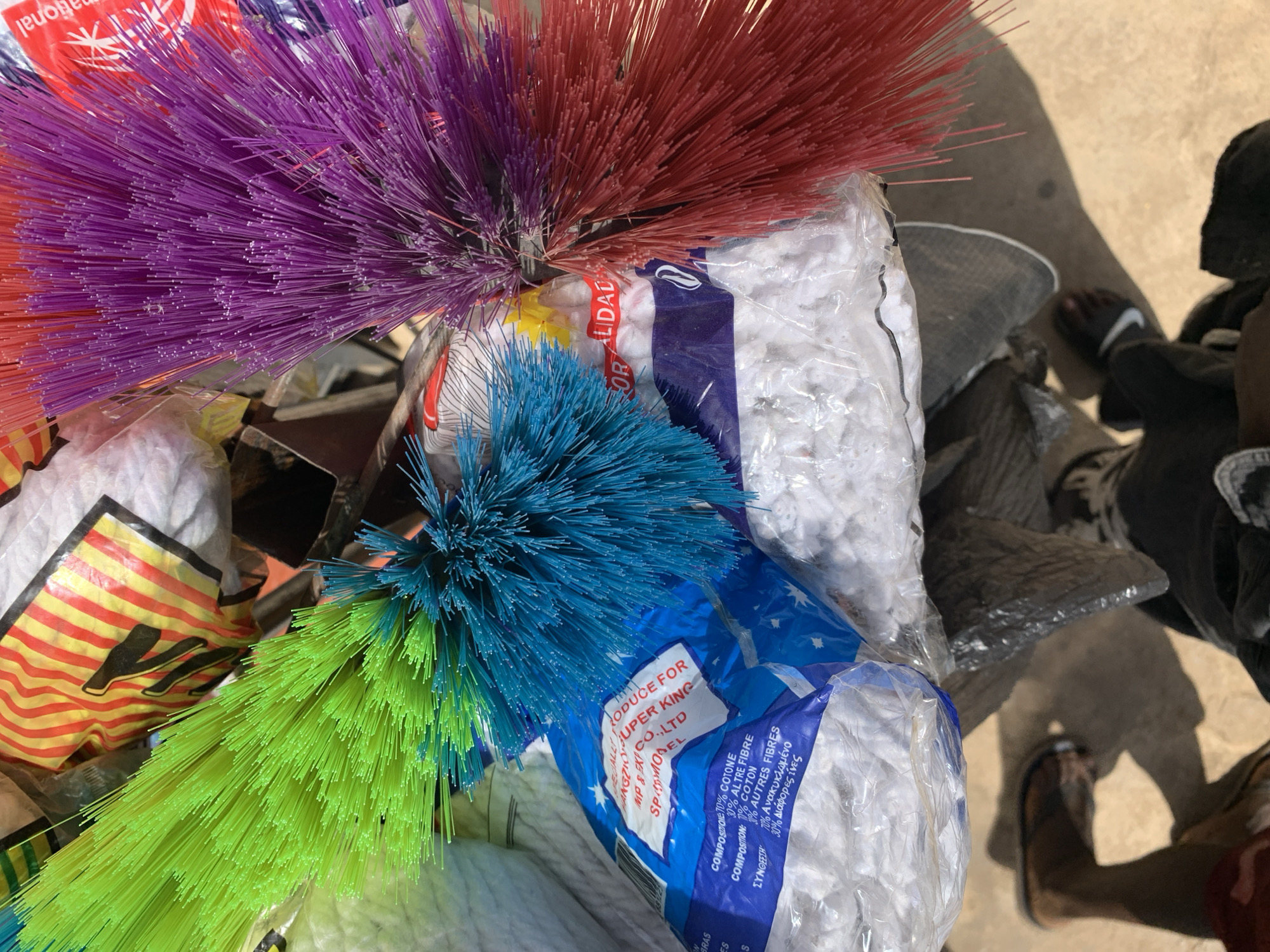 The image shows a close-up of a collection of colorful cleaning brushes and mops in plastic packaging. The brushes are purple, red, blue, and green. The mop packaging is labeled in English and possibly Greek, mentioning "SUPER KING Co. LTD" and composition details (cotton and other fibers). The presence of shadows and a partially visible person wearing sandals and dark clothing suggests the setting is outdoors, likely in direct sunlight. Although the description mentions the city of Jos, Nigeria, this is not discernible from the provided image. Therefore, any association with a specific location would need additional context.