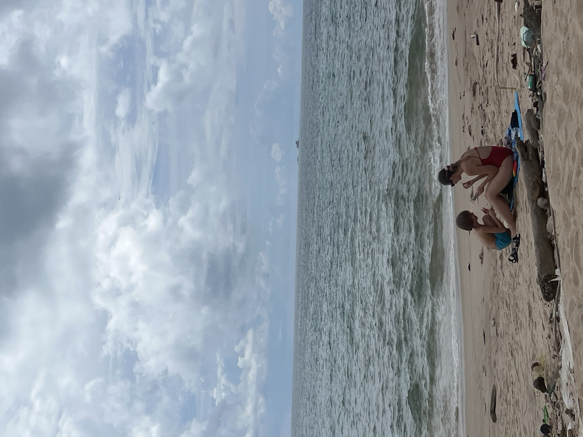 A woman and a child sit on a sandy beach in Choeng Thale, Thailand, facing the sea. The woman is wearing a red swimsuit and sunglasses, and the child is wearing blue swim trunks. They are both looking down, and it appears they are interacting with something on the sand. The sea is choppy with small waves, and the sky is cloudy with some patches of blue. The beach is littered with driftwood, plastic bottles, and other debris. In the distance, there are a few small islands.