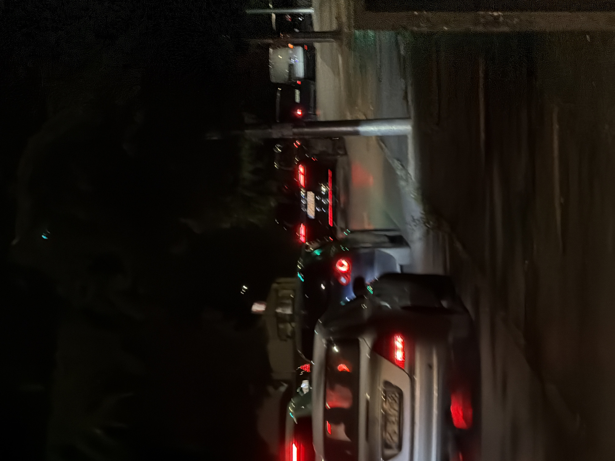 The image depicts a night scene in Podgorica, Montenegro, with a line of cars visible on a road. The vehicles appear to be stopped or moving slowly. The location appears to be an area with overhead structures or bridges, possibly along a main thoroughfare. Illumination is provided by vehicle headlights and taillights. The environment is dark.
