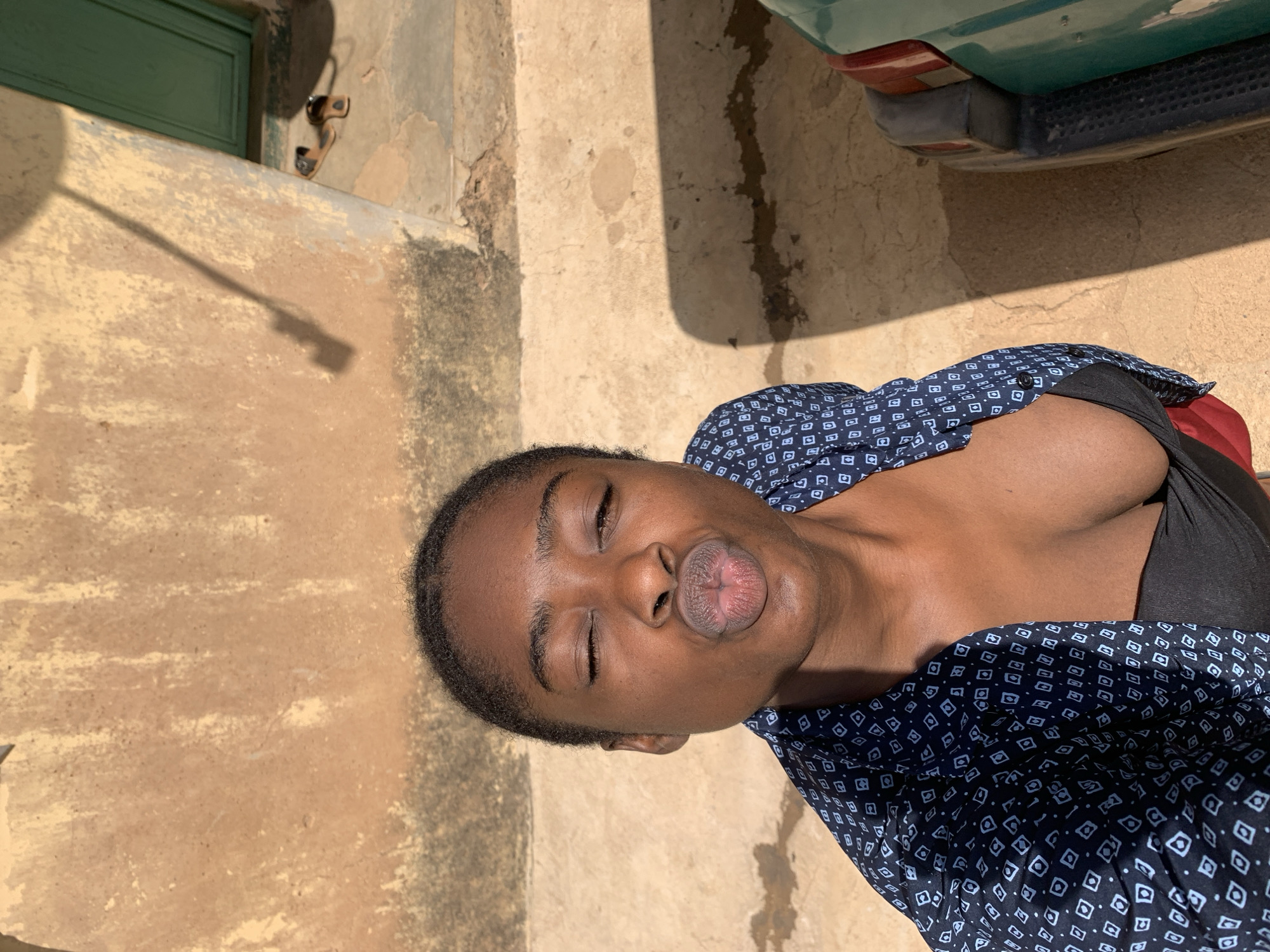 The image shows a woman with dark skin pursing her lips in a kiss expression. She is wearing a dark shirt or top underneath a blue and white patterned button-down shirt. The background consists of an exterior wall with patches of discoloration, likely from age or weather. A portion of a green vehicle is visible in the upper right corner of the frame. Based on the information given the location is likely in the city of Rafin Par, Nigeria.