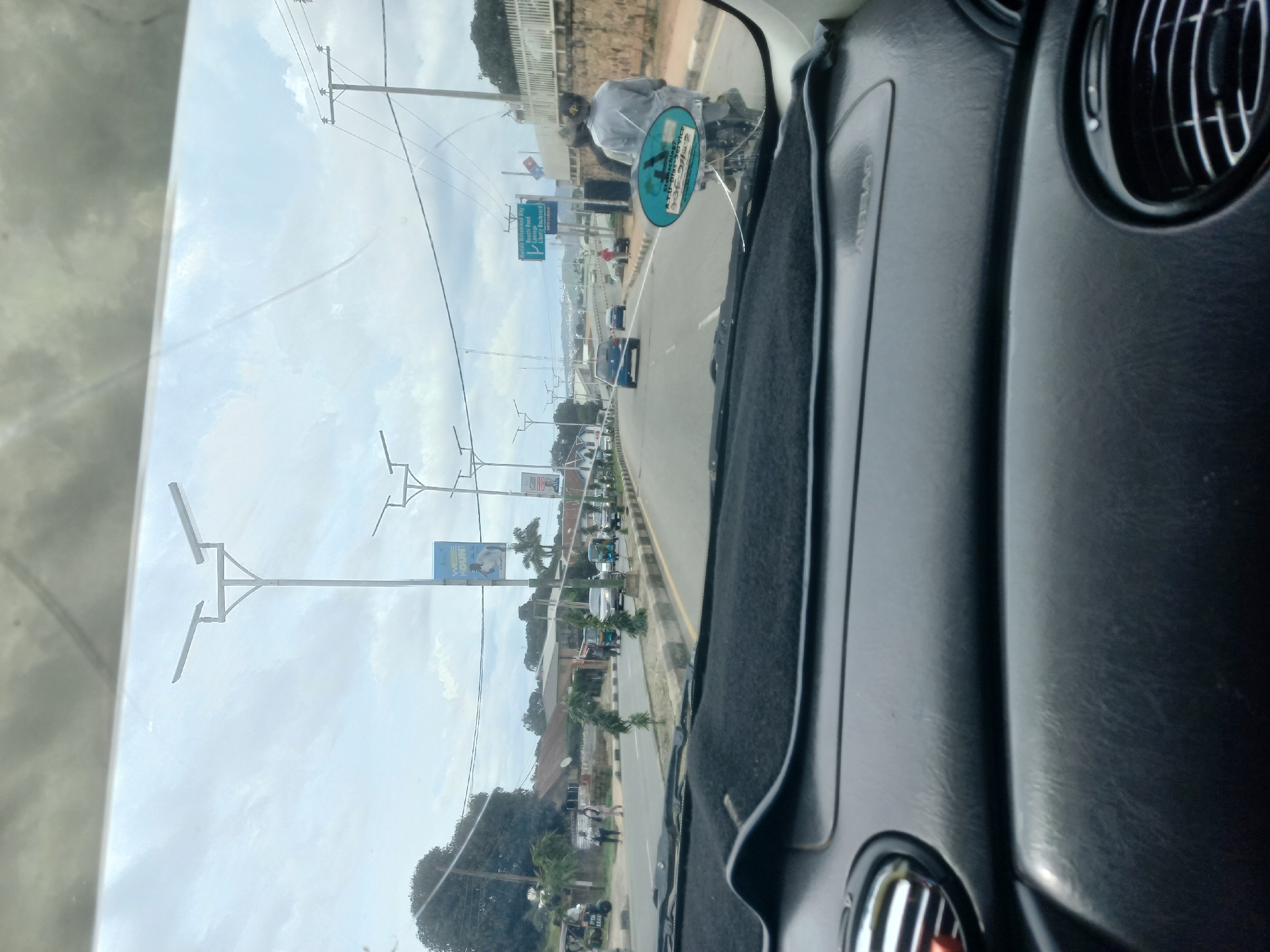 The image, taken from the interior of a vehicle looking forward through the windshield, depicts a street scene in Jos, Nigeria, on a cloudy day. The road is paved and appears to be a multi-lane thoroughfare. Several vehicles are visible, including cars and what appear to be "Keke" (motorcycle taxis). Pedestrians are present on the sidewalks and crossing the street. Streetlights with solar panels line the road, and power lines are strung overhead. Buildings of varying styles are visible along the roadside, some with corrugated metal roofs. There are also signs and billboards, one of which appears to be a directional sign for "Harold Mohammed Way," "Bauchi Road," and "Liberty Boulevard." A person on a motorcycle, wearing a light-colored outfit and a dark cap, is in the foreground on the right, with an oval sticker on the back of the motorcycle. The interior of the vehicle shows a dashboard with an "AIRBAG" label. The environment appears to be urban, with a mix of infrastructure and vegetation.