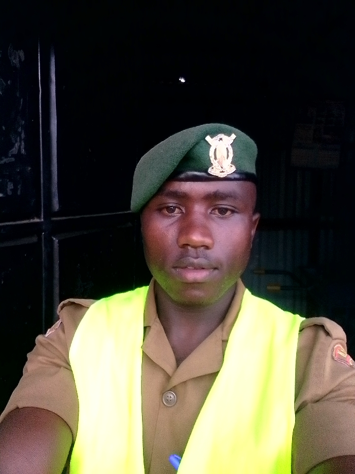A man in a tan uniform and a green beret with a badge stands facing the camera. He wears a bright yellow safety vest. The background is a dark doorway or entrance. The man's skin tone is dark, and he has a serious expression. A pen is visible near the bottom of the image. He appears to be a security guard or a member of a disciplined force.