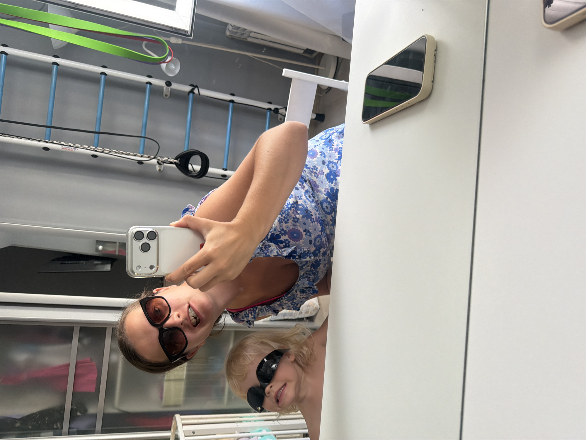 In Sochi, Russia, a woman and a young child are reflected in a mirror, posing for a selfie. The woman, wearing large, dark sunglasses and a blue floral dress, holds a smartphone. The child, also sporting dark sunglasses, sits beside her. The background reveals a home gymnasium with a wall-mounted ladder and resistance bands. A white table occupies the foreground.