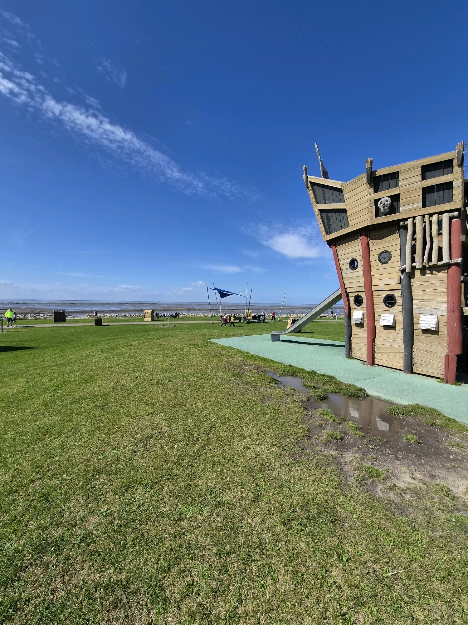 The image depicts a playground area in Dorum, Germany, featuring a large wooden pirate ship-themed play structure with a slide. The structure is located on a green, grassy lawn. In the background, the North Sea can be seen with a beach and a walkway. Several people are visible in the distance, some walking and others appearing to be seated, possibly at beach chairs. The sky is bright blue with scattered white clouds. The foreground shows a patch of wet, muddy ground adjacent to the green playground surface, indicating recent rainfall or water accumulation.