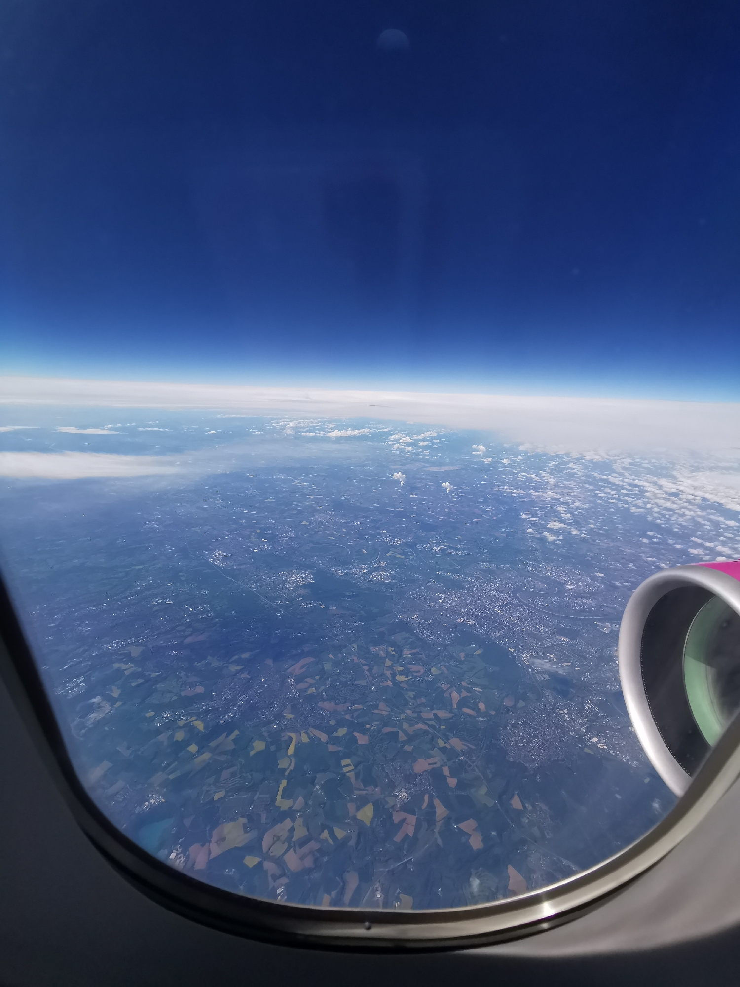 The image shows a view from an airplane window. The sky is blue above a layer of clouds near the horizon. Below the clouds, the land is visible with a patchwork of fields and urban areas. Part of the airplane's engine is visible in the lower right corner of the window, with pink detailing. The geographic location is undetermined, and is not verifiable as the city of Varna, Bulgaria. There are no visible people or interactions. The scene depicts a view from a high altitude.