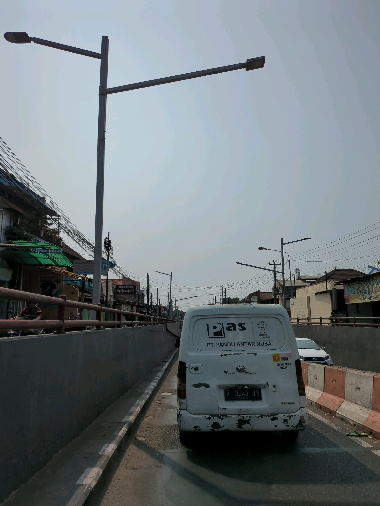 The image depicts a street scene in Purbayan, Indonesia, characterized by a raised roadway with concrete barriers on either side. The sky is overcast and hazy. In the foreground, a white van with the logo "PT. PANDU ANTAR NUSA" is visible, facing away from the camera. The van has a license plate reading "R 8043 IR". To the left of the roadway, a concrete retaining wall separates the street from buildings and dense overhead wiring. A motorcycle rider is partially visible behind a reddish-brown metal railing on the left. Further down the street, more buildings and streetlights are visible. On the right side of the roadway, another vehicle, a white car, is partially visible. Orange and white concrete barriers line the right side of the street. A single flip-flop is lying on the edge of the roadway to the right. The overall impression is a busy urban environment with overhead infrastructure.