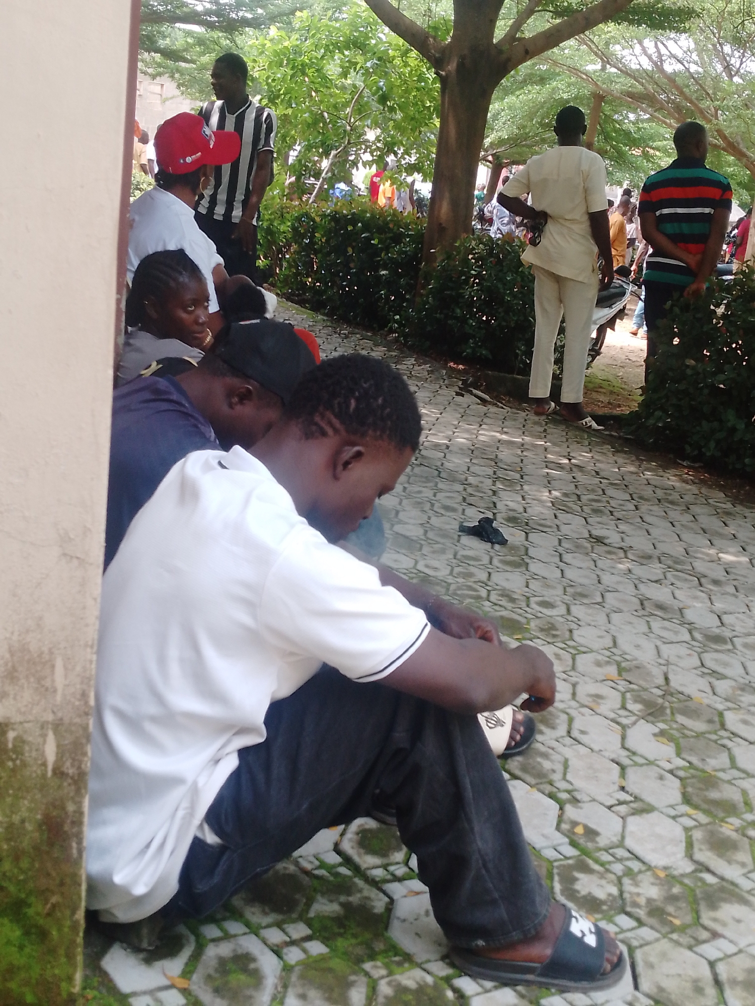 In Akwanga, Nigeria, a group of individuals are gathered on a paved walkway. In the foreground, a person wearing a white t-shirt and black pants sits on the ground, facing away from the camera. To their left, another person in a blue shirt and black baseball cap is also seated. Further back, a person in a red baseball cap and white t-shirt is visible. Behind them, several other individuals are standing and interacting. The environment appears to be an outdoor area with trees and lush green foliage. The walkway is made of hexagonal paving stones.