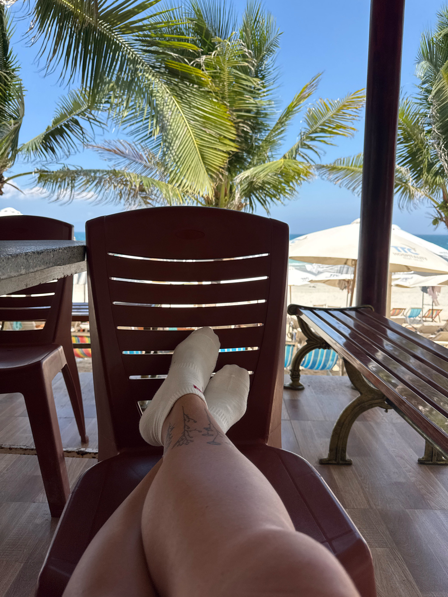 The image depicts a first-person perspective of someone relaxing on a brown plastic chair. The individual's legs are crossed, and they are wearing white socks, one of which has a subtle tattoo of a plant-like vine on the ankle. The background reveals a beach scene with palm trees swaying under a clear blue sky. White umbrellas and beach chairs are visible on the sandy beach, and the ocean can be seen in the distance. The setting appears to be a covered area, possibly a restaurant or bar, as indicated by the presence of tables and chairs. The overall ambiance suggests a tropical vacation or a leisurely day by the sea. The location is identified as Dong Ninh Hoa, Vietnam.