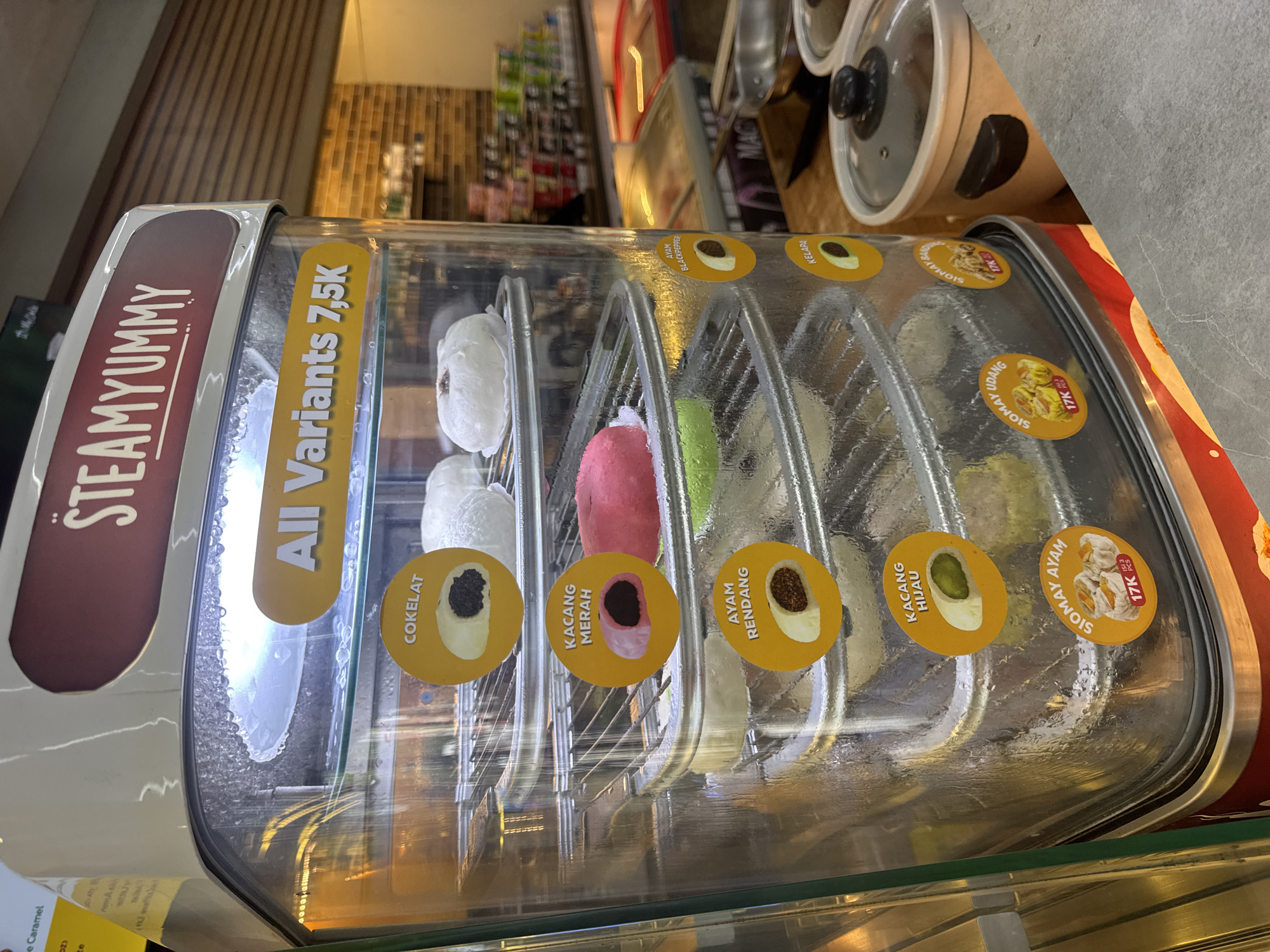 The image shows a steaming display case for steamed buns and siomai, branded as "Steamyummy." The display case is filled with various flavors of steamed buns, including "Cokelat" (chocolate), "Kacang Merah" (red bean), "Ayam Rendang" (rendang chicken), and what appear to be green-colored buns likely representing pandan or avocado. Above the buns, a yellow sign reads "All Variants 7,5K." On the side of the display case, there are additional labels for "Ayam Blackpepper," "Kelapa" (coconut), "Siomay Ayam" (chicken siomai), "Siomay Udang" (shrimp siomai), and "Siomay Bandeng" (milkfish siomai), some with prices indicated as "17K." The display case is positioned in what appears to be a food stall or small eatery, with a blurred background showing more food displays, shelving with products, and brick wall elements. The foreground shows a concrete countertop and a ceramic pot with a lid. The overall setting suggests a casual food establishment in a tropical climate, likely Kuta Selatan, Indonesia, given the context provided.