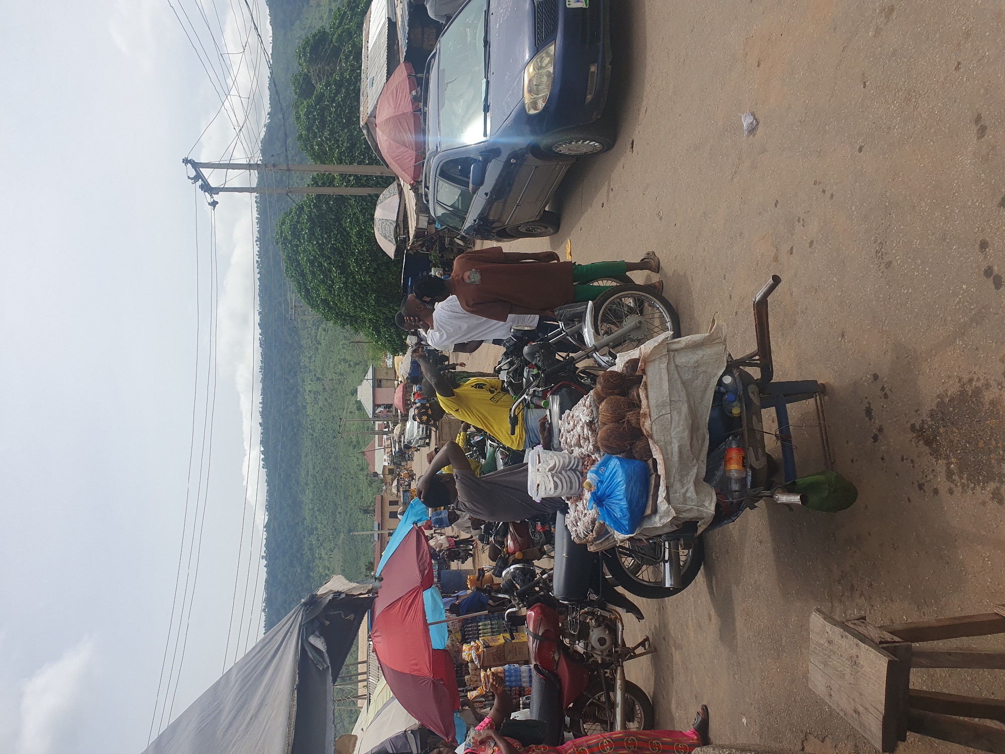 The image depicts a bustling street scene in Lokoja, Nigeria. The focal point is a motorcycle cart loaded with coconuts and other goods, likely for sale. People are standing around the cart, possibly engaging in transactions. Other motorcycles are parked nearby, some with similar displays of merchandise. A blue car is parked close to the street with at least one red umbrella visible in the background providing shade to sellers and goods. Overhead, power lines traverse the scene. In the background, a lush, green hill provides a natural backdrop. The lighting is bright, suggesting a sunny day. The road surface appears to be asphalt and is partially dirty. The image showcases typical urban street life in Lokoja.