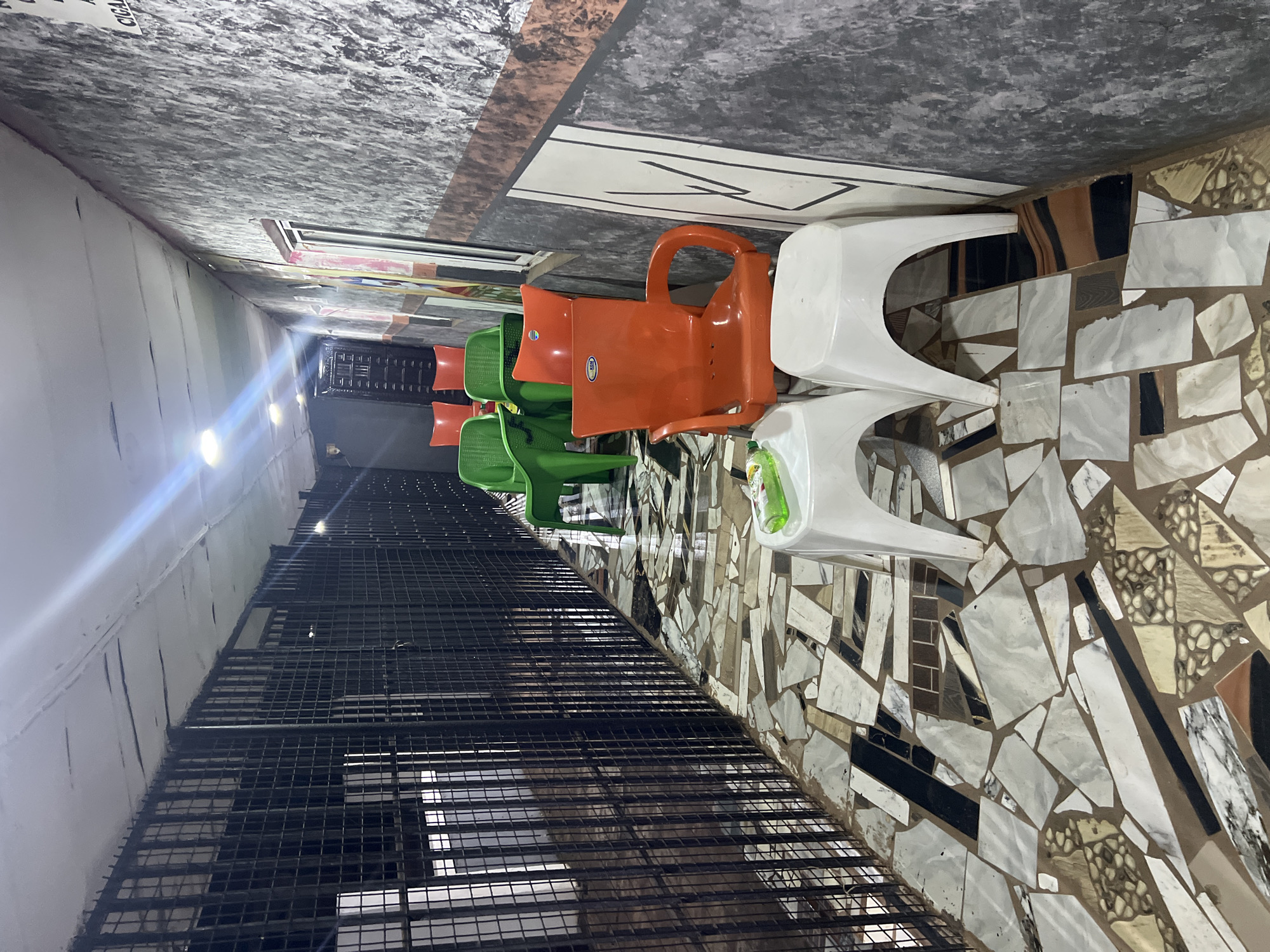 The image depicts an outdoor walkway with a tiled floor and a barred enclosure on the left. Several plastic chairs are arranged along the walkway, with some in green and others in orange. Two white plastic stools are placed in the foreground. The walls are textured and appear to be made of concrete or stone. A sign with "CIGA" visible is attached to the right wall. The scene is lit by artificial lights, and the walkway appears to be open to the elements at the far end. The location is identified as Nkpor, Nigeria.