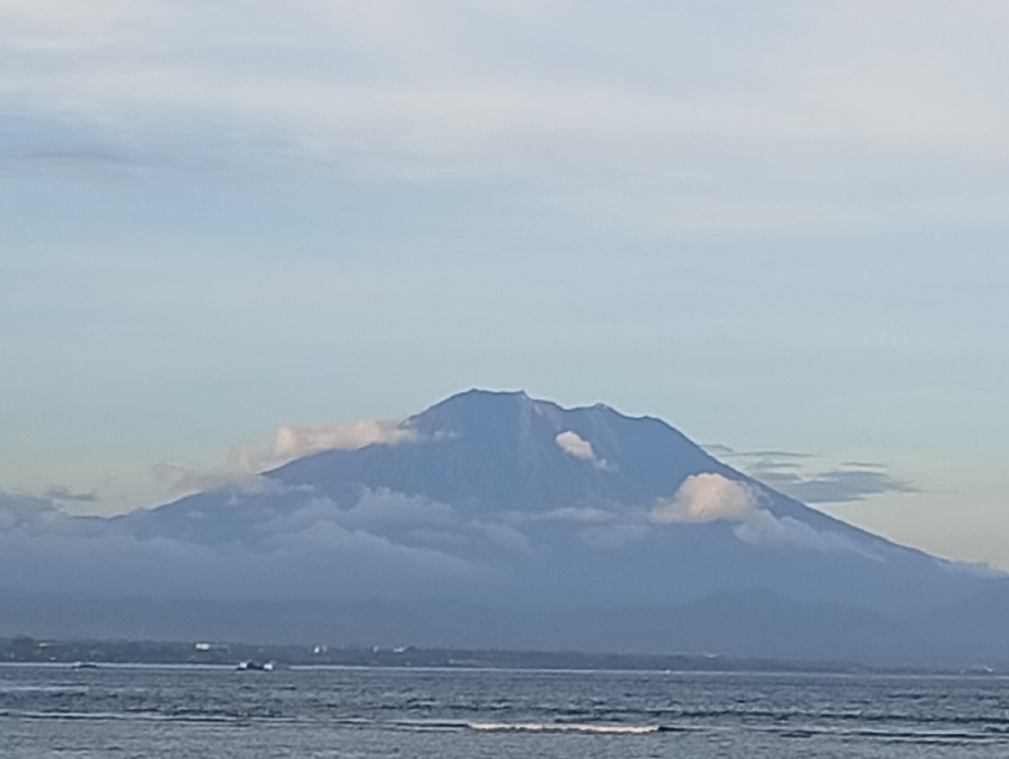 The image presents a serene landscape dominated by a majestic mountain rising behind a calm body of water. In the foreground, the sea exhibits subtle ripples and gentle waves, with a few indistinct, dark shapes that appear to be boats or small vessels navigating the distant waters. A faint, low-lying strip of land or coastline can be discerned between the water and the mountain, featuring some barely visible structures.
The central and most prominent subject is a large, imposing mountain, rendered in shades of blue-grey, indicating its distance and the atmospheric haze. Its rugged peaks are partially enveloped and adorned by soft, white clouds, particularly along its slopes and crest. The sky above is a clear, pale blue, suggesting the soft light of either early morning or late afternoon. The overall scene conveys a sense of tranquility and natural grandeur. Based on the provided location context of Denpasar, Indonesia, the mountain is identifiable as Mount Agung in Bali. No text is visible in the media file.