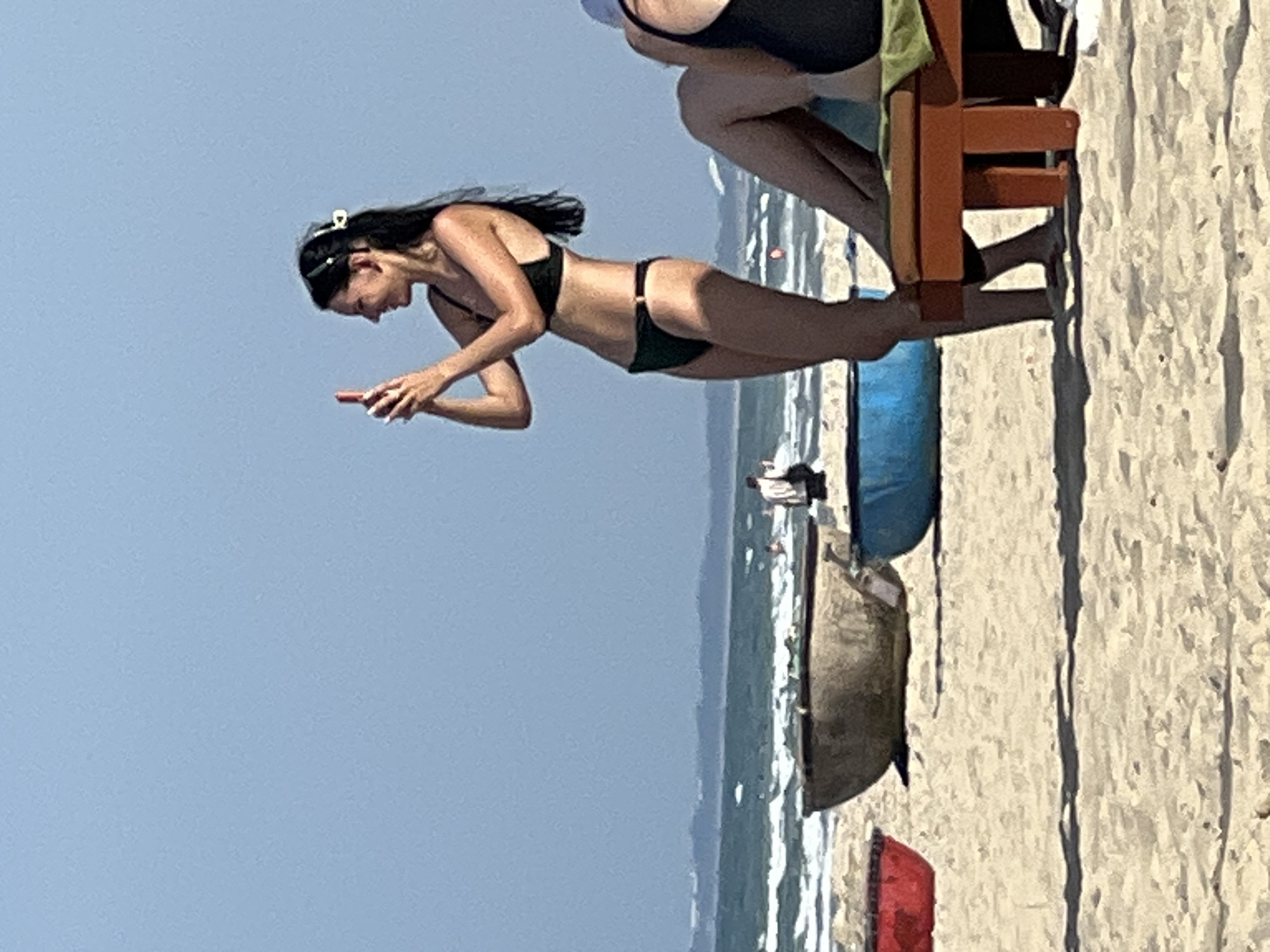 The image shows a beach scene in Đà Nẵng, Vietnam, with the ocean in the background and sand in the foreground. A young woman in a black bikini is standing on the sand, holding up a mobile phone to take a picture. She has dark hair, pulled back with a white clip. To her right, partially visible, is a person sitting on an orange beach chair with a green towel draped over it. In the mid-ground, there are three boats on the sand: a red one on the left, a weathered brown boat in the center, and a blue boat on the right. The sky is clear and blue.