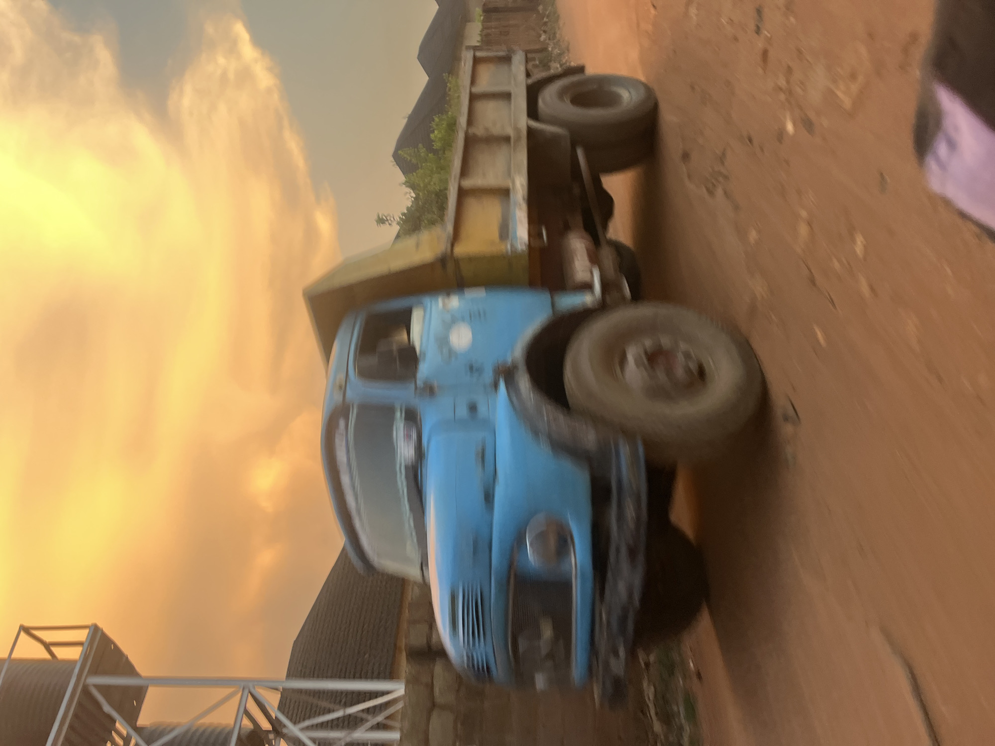 The image shows a blue and yellow dump truck on a dirt road. The truck is facing the viewer. The sky in the background is a mix of orange and yellow. There is a partially visible metal structure in the lower left corner. The photo's angle is distorted and the scene appears to be in Abuja, Nigeria.
