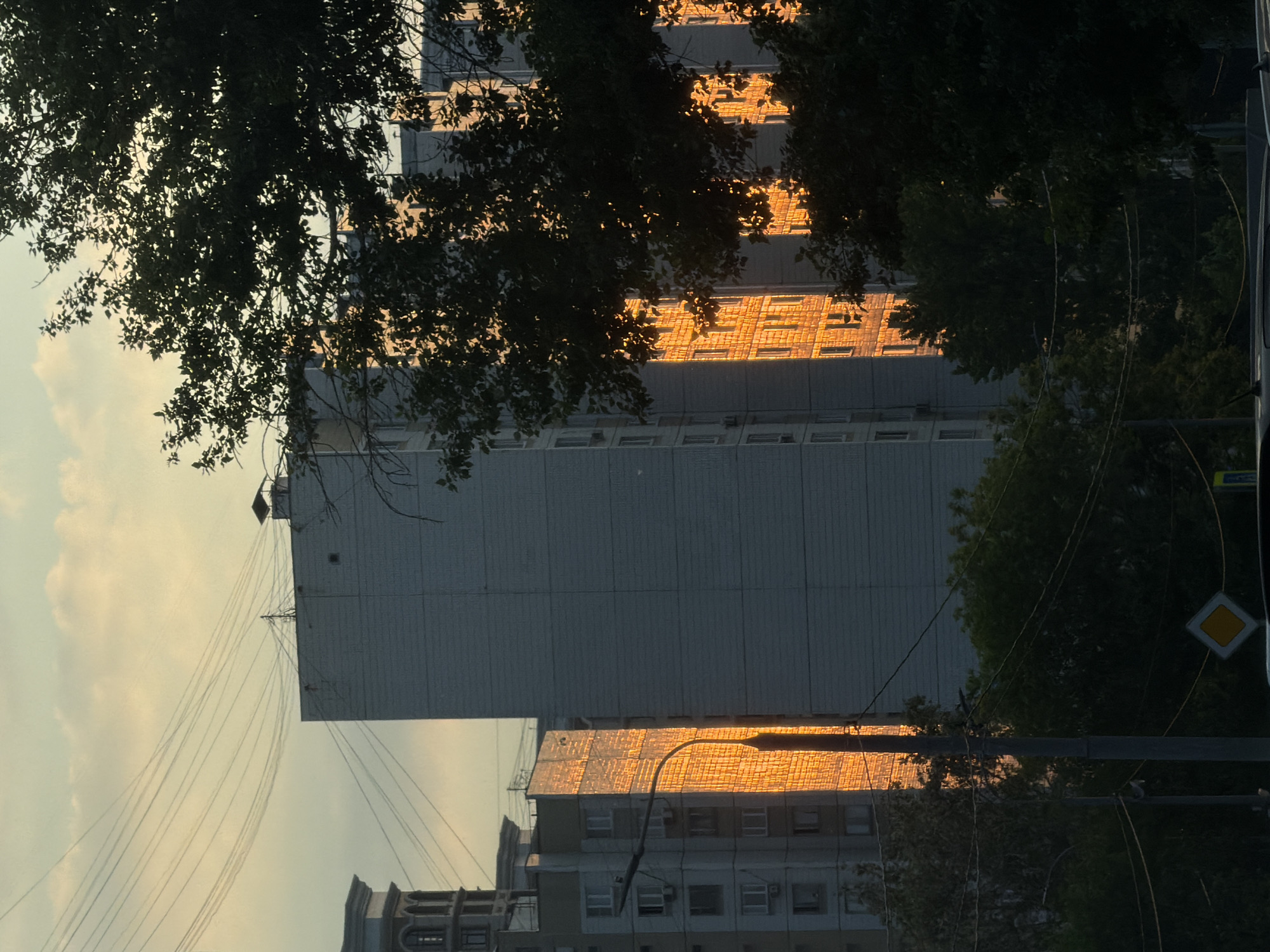 The image depicts a late afternoon or early evening view of a residential area, likely in Moscow, Russia, given the architectural style of the buildings and the presence of overhead electrical wires. The sky is a pale, hazy yellow, indicative of a sunset or sunrise, with scattered clouds. The main subjects are several multi-story apartment buildings. One building in the center, constructed with light-colored bricks, is dramatically illuminated by the golden sunlight, highlighting its windows and brickwork. To its left, another, slightly shorter building with a similar facade also catches the golden light, with a lamppost in front of it. To the right, a large, dark green tree with dense foliage partially obscures the view of the buildings. The foreground is dominated by the dark silhouette of more trees and the tops of vehicles, suggesting a street-level perspective. Overhead, numerous electrical wires crisscross the sky, a common feature in urban Russian landscapes. A yellow diamond-shaped traffic sign is visible on a pole in the lower part of the frame, further contributing to the urban setting. No people are visible in the image.