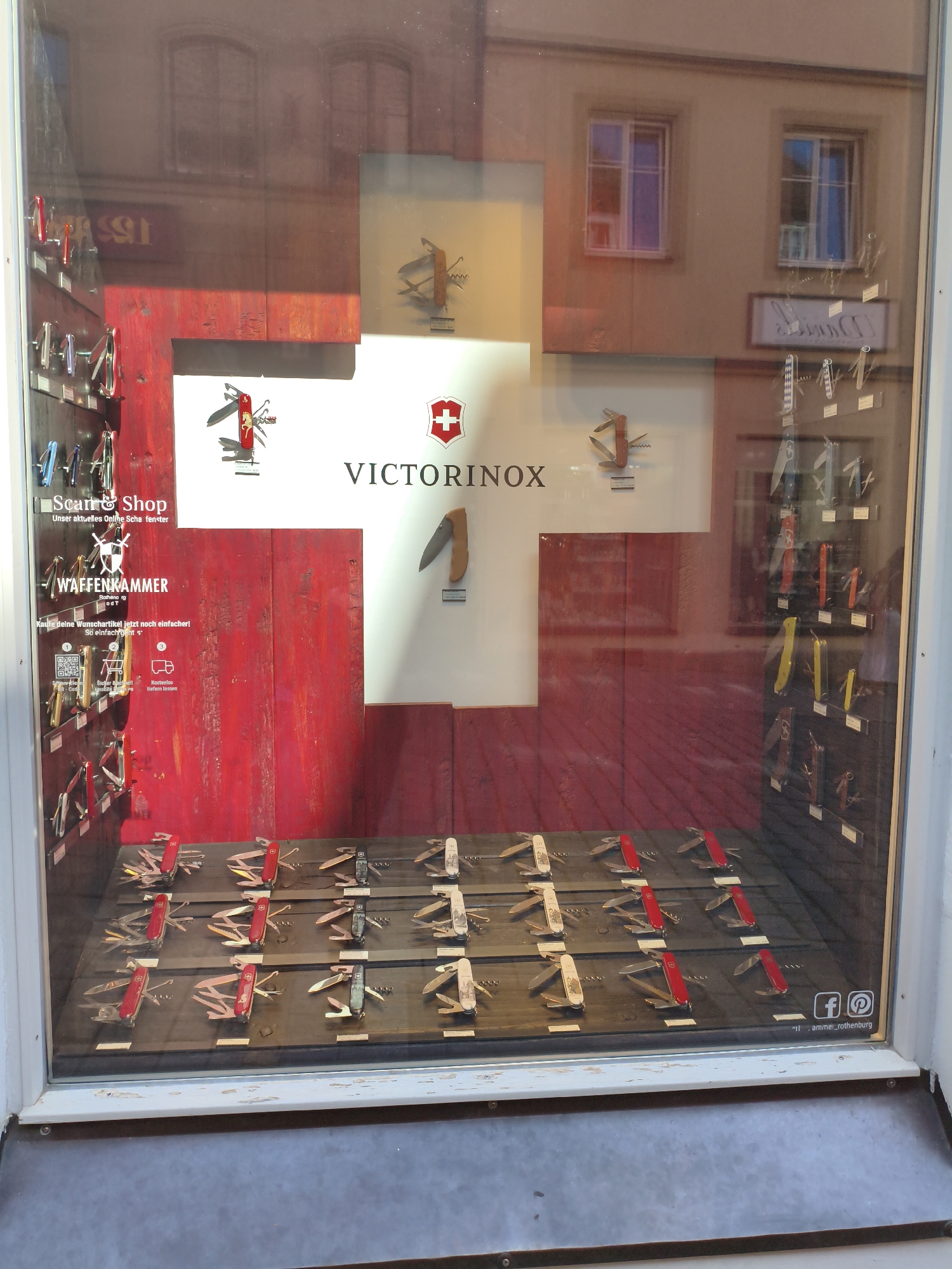 The image displays a shop window in Rothenburg ob der Tauber, Germany, showcasing an assortment of Victorinox Swiss Army knives. The display is set against a large red cross emblem, a nod to the Swiss flag and the brand's origin. Various knives are arranged on shelves and on a wooden tiered display in the foreground. Some knives are mounted on a red wooden structure on the left, accompanied by text that includes "Scan & Shop" and "WAFFENKAMMER." The background reflection in the glass shows buildings with windows. Social media icons for Facebook and Instagram, along with the handle "armier_rothenburg," are visible in the lower right corner of the window.