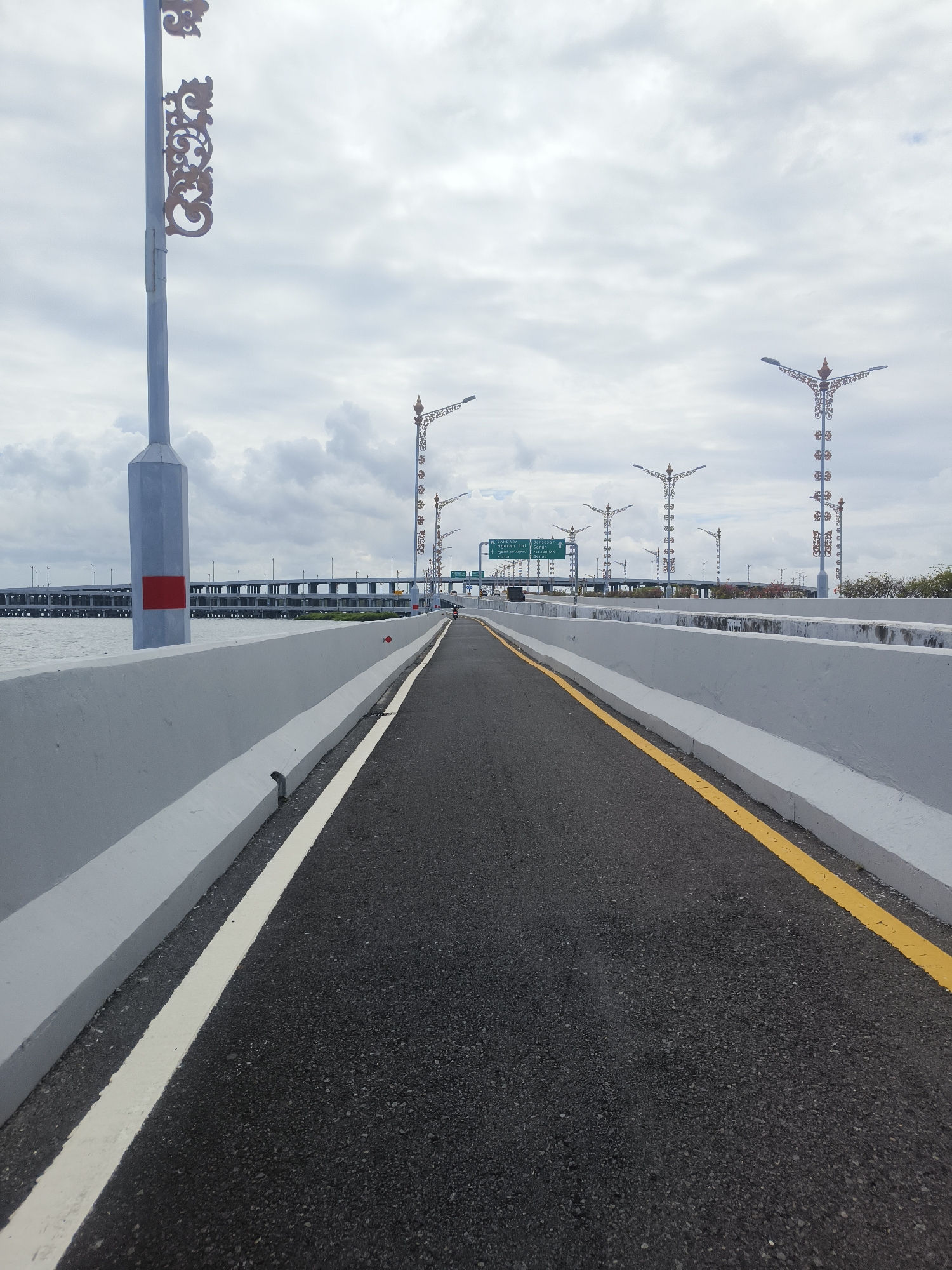 The image depicts a perspective shot of a modern road on a bridge, likely in Kuta Selatan, Indonesia, given the presence of ornate street lamps that are characteristic of the region. The road surface is asphalt, marked with a white line on the left and a yellow line on the right, delineating lanes. A low concrete barrier runs along the sides of the road. In the background, a complex network of elevated roads and structures suggests ongoing or completed infrastructure development. The sky is overcast with scattered clouds. Several decorative street lamps with intricate designs are visible along the bridge. In the far distance, a few vehicles and a person on a motorcycle are present. The water body on the left side of the bridge indicates it spans over water.