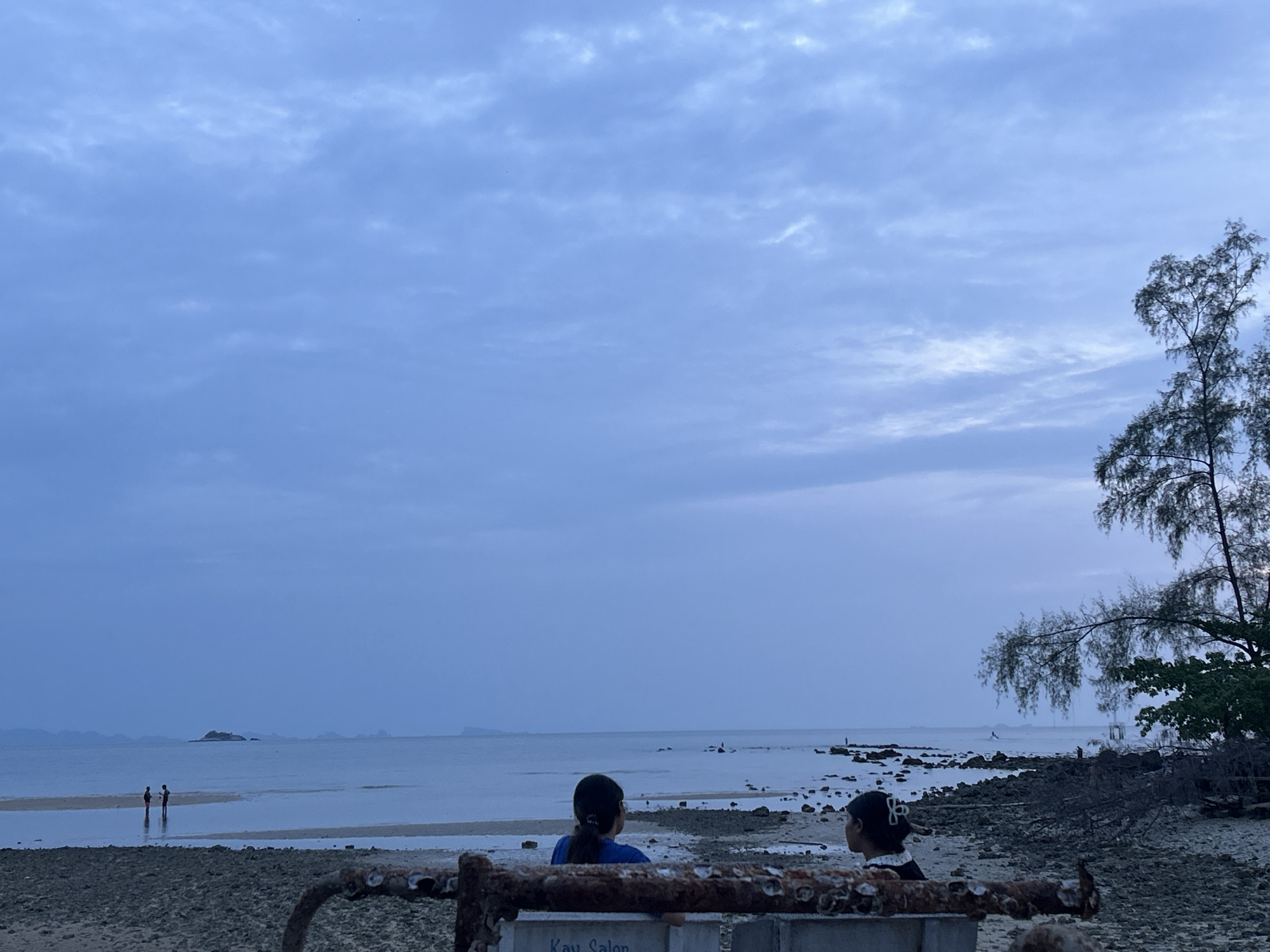 The image captures a coastal scene in ตำบลเกาะพะงัน, Thailand, under a predominantly cloudy sky. Two individuals are seated in the foreground, their backs to the camera. In the background, two figures are wading in the shallow water. The environment includes trees to the right and a small island in the distance. The foreground contains a rusty structure, and the beach is rocky.