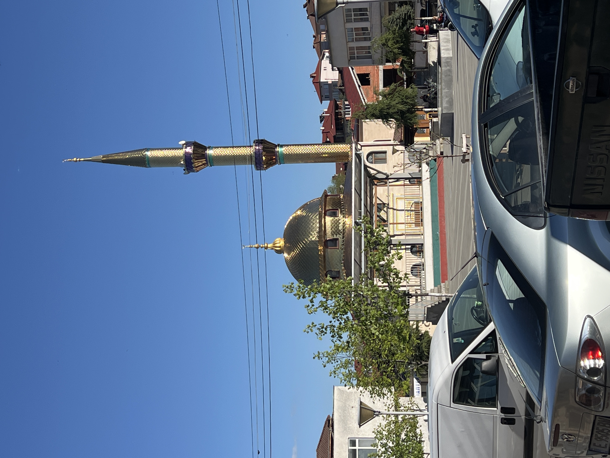 The image shows the Golden Mosque in the city of Breznitsa, Bulgaria. The mosque's golden dome and minaret are prominently visible against a clear blue sky. Power lines run across the top left portion of the frame. Buildings of varying heights and colors surround the mosque. Parked cars, including a Nissan, are visible in the foreground. Green trees and vegetation are also present. The scene appears to be captured during the day, likely in the morning or afternoon, given the light.