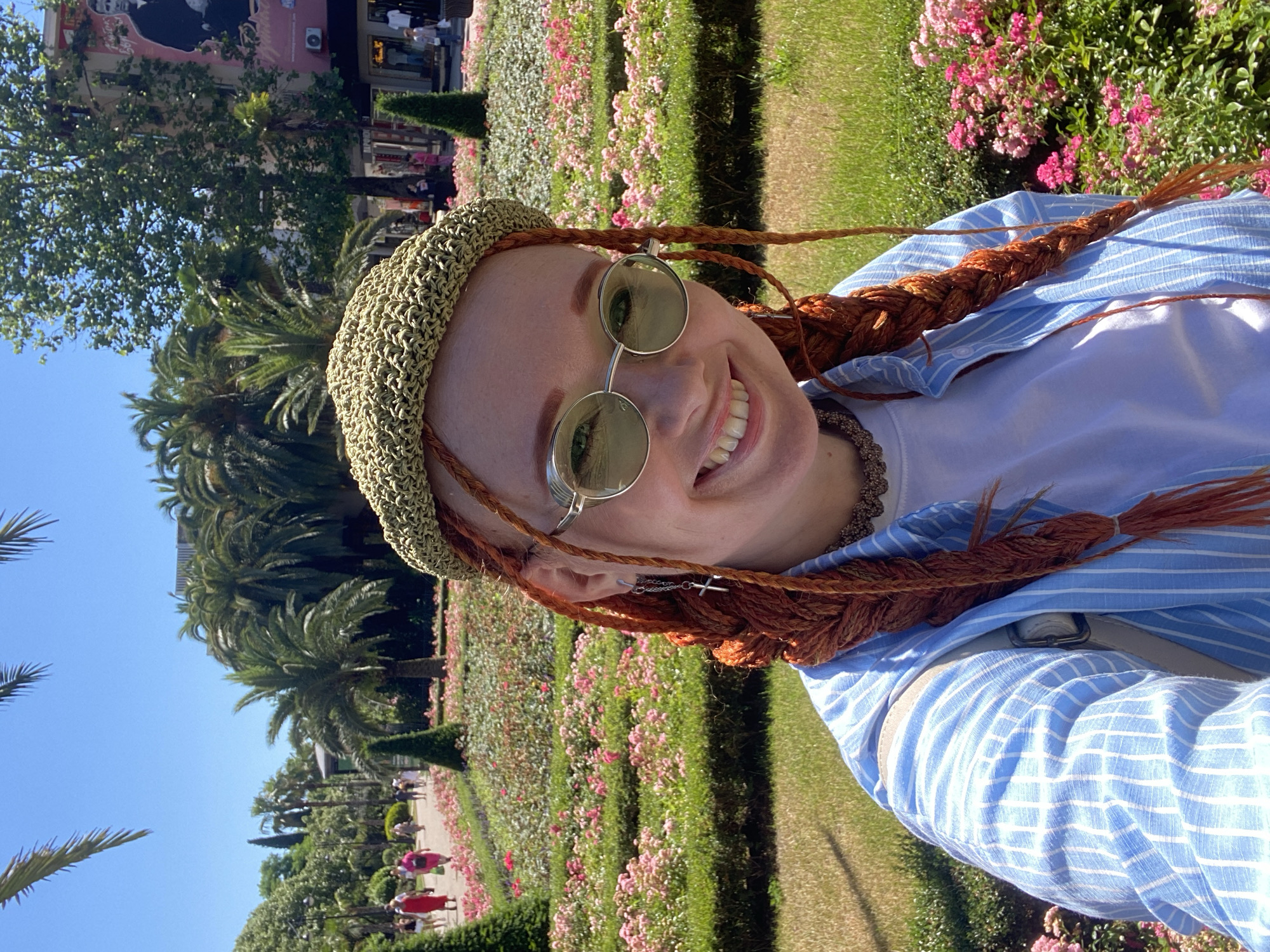 A young woman with long, braided, orange hair and a green crocheted hat takes a selfie in front of a park with abundant pink flowers, palm trees, and a clear blue sky. She is wearing round, green-tinted sunglasses, a white t-shirt, and a light blue and white striped button-down shirt. A cross earring hangs from her left ear. In the background, people walk along a path, and buildings with signage are visible. The scene appears to be outdoors in a public park on a sunny day, possibly in the city of Sochi, Russia, given the tropical foliage.
