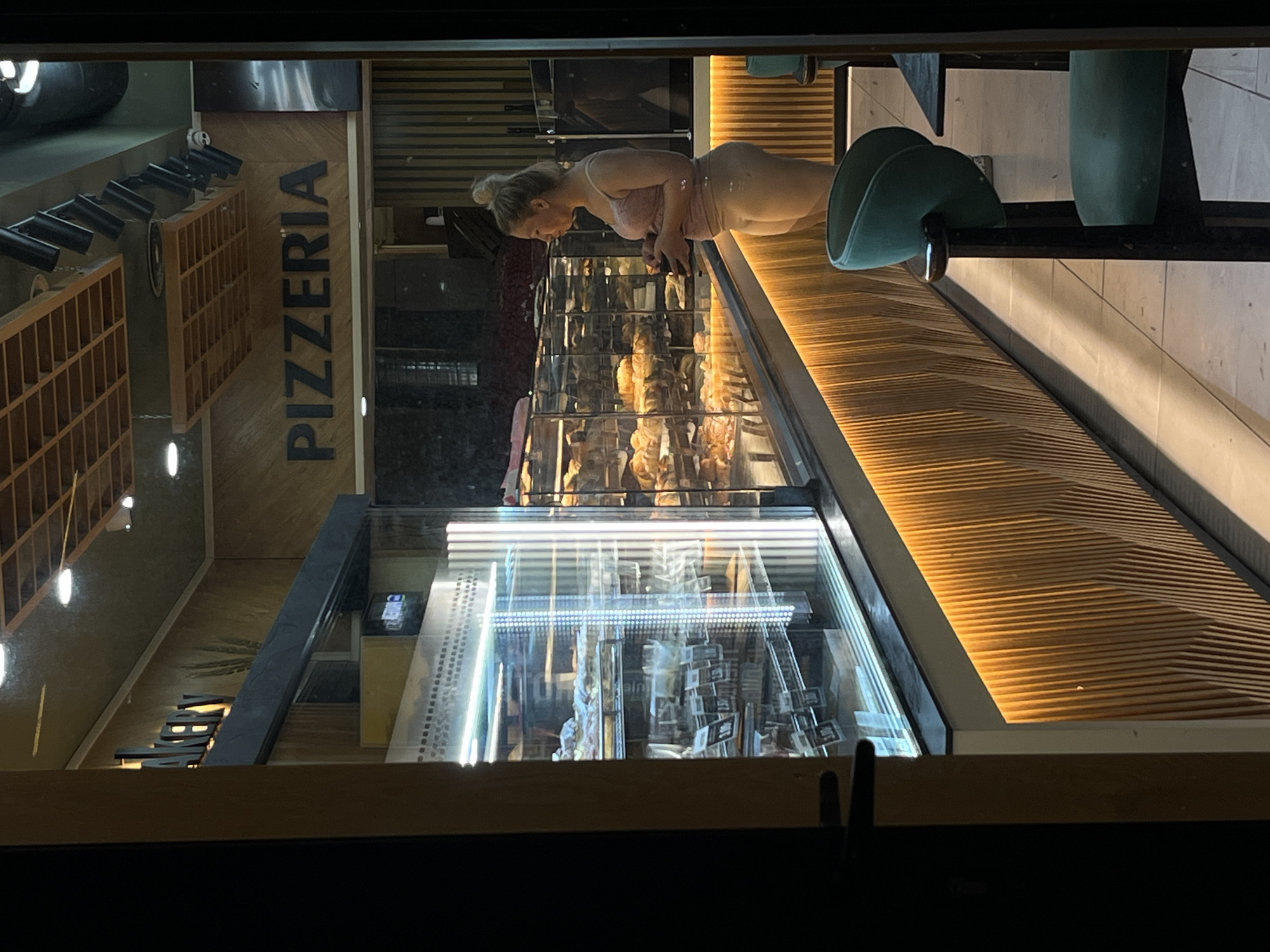 The image depicts the interior of a pizzeria in Budva, Montenegro, viewed through a storefront window at night. A young woman with blonde hair pulled into a bun stands inside the establishment, facing away from the camera and towards a refrigerated display case filled with food items. She is wearing a light pink top and tan shorts. The display case is brightly lit, illuminating various pastries and possibly sandwiches within. Behind the woman, the word "PIZZERIA" is prominently displayed on the wall in large, dark letters. The pizzeria features a modern design with wooden paneling and warm, integrated lighting along the base of the display counter. A dark green chair is visible in the foreground to the right of the image.