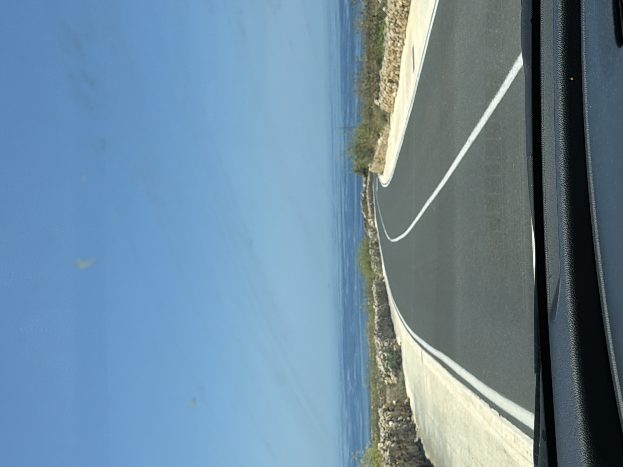 The image depicts a road curving towards the right, bordered by a dry stone wall on the left and vegetation on the right. The road has a white dividing line. Beyond the wall and vegetation, the blue sea stretches out, meeting the clear blue sky. The foreground shows the dashboard of a vehicle. The scene is likely located in a coastal area, possibly along the road leading to Ta' Ġawhar in Saint Lawrence, Malta, which is known for its scenic coastal routes. There are no people or animals visible in the image. The overall impression is of a bright, sunny day with clear visibility.