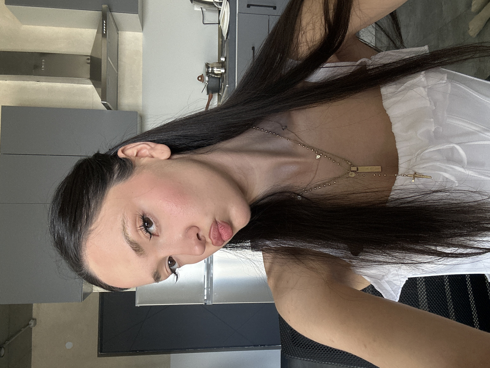 A selfie of a woman with long, dark hair wearing a white, ruffled top and a gold necklace with a cross pendant, set against a kitchen background. The kitchen features gray cabinets, a stainless steel refrigerator, and a cooker hood. The location is likely indoors. There is no information that allows me to confidently identify that the photo was taken in Zovuni, Armenia.
