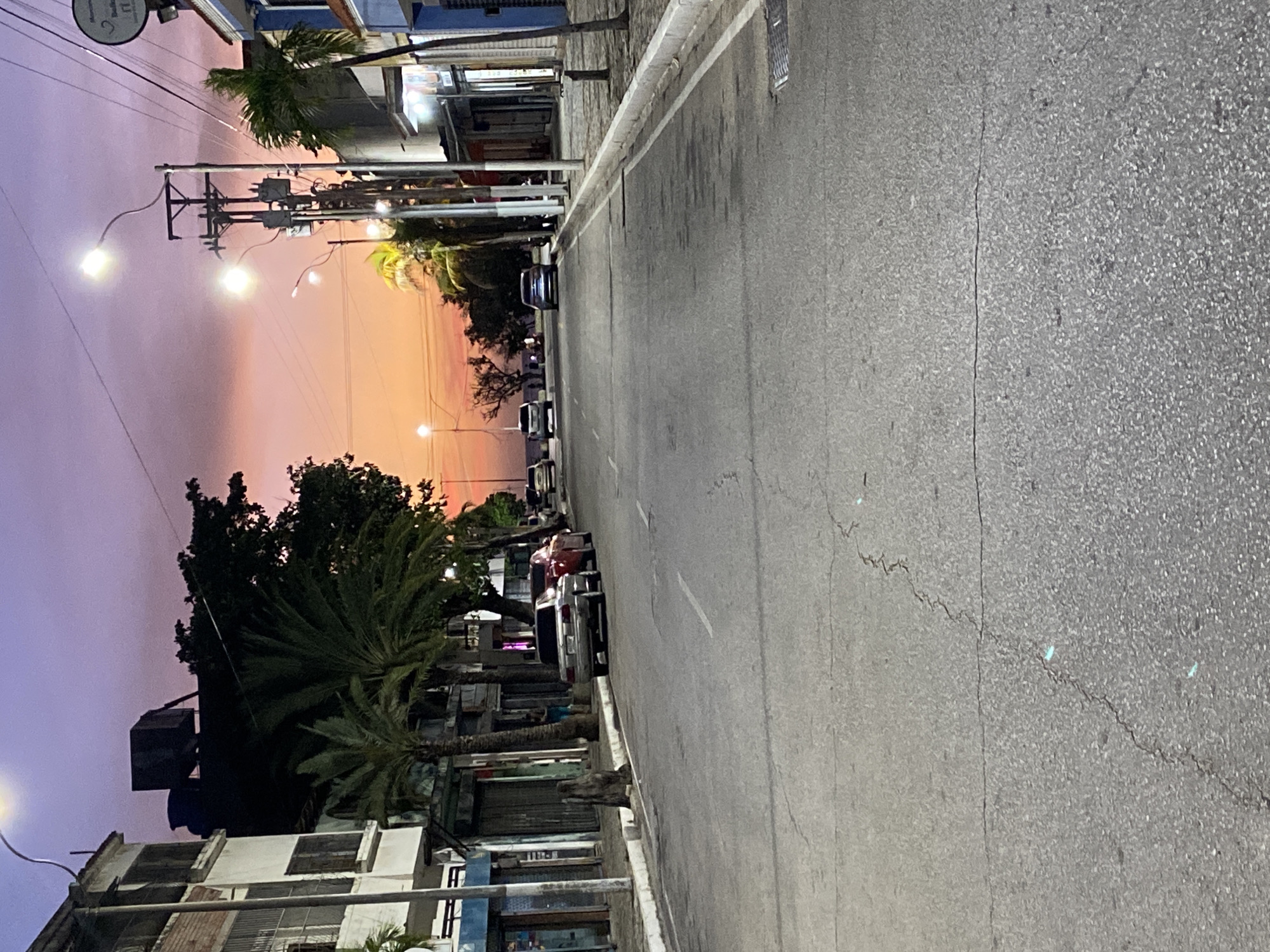 This image captures a street in Juan Griego, Venezuela, likely at dusk or dawn. The sky displays a gradient of orange and purple hues. On either side of the road, buildings line the street, with palm trees interspersed among them. Cars are parked along the side of the road. Streetlights illuminate the scene. The road itself is asphalt, with a dividing line and some visible cracks.