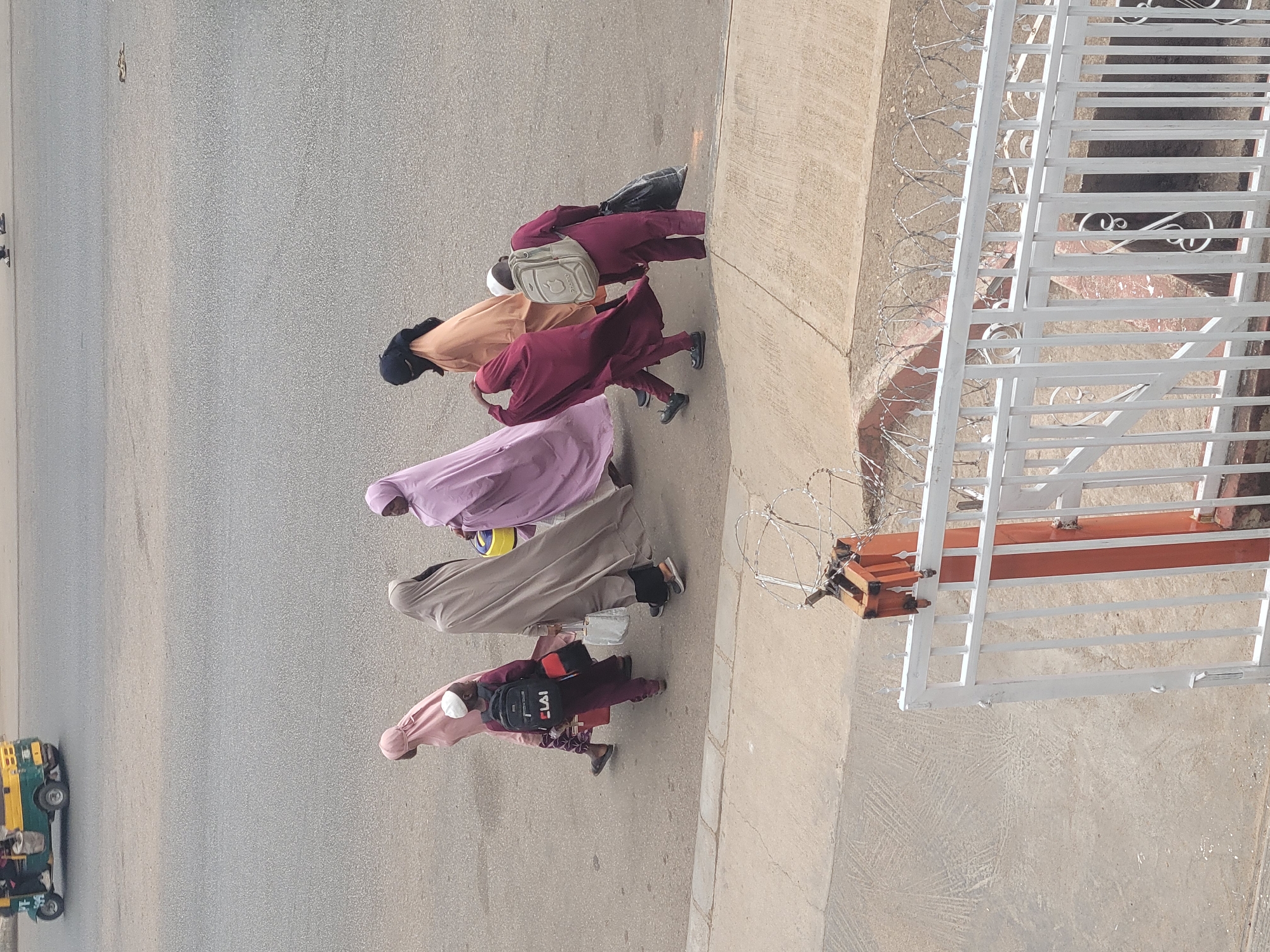 Five women, wearing colorful hijabs and outfits, are walking in a line down a street, away from the camera. They are walking on the sidewalk. A yellow vehicle is also on the road. A metal gate and barbed wire are on the right side of the image. The location is Jos, Nigeria.