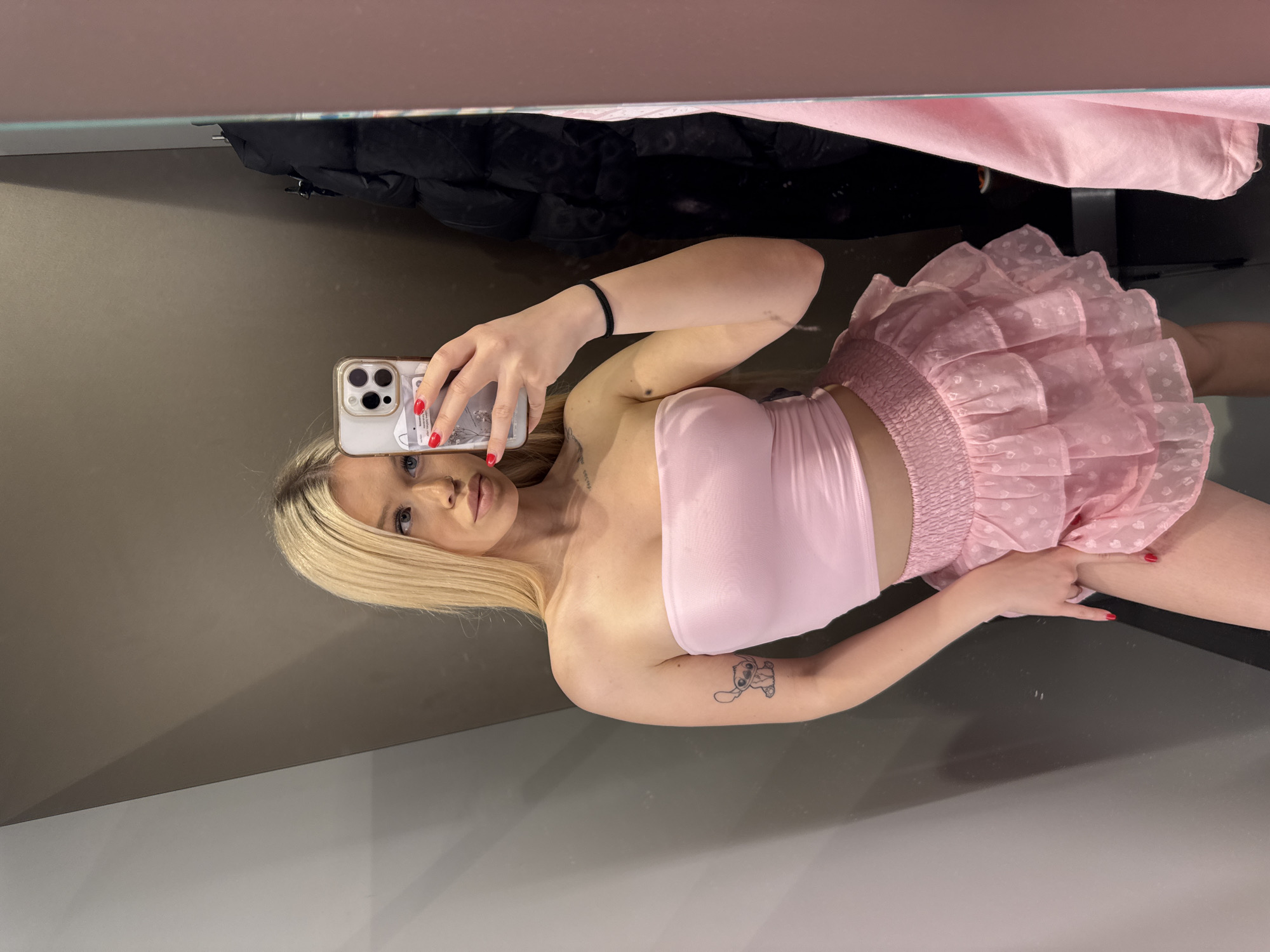 A young woman with blonde hair, wearing a pink strapless top and a pink ruffled skirt, takes a selfie in a fitting room. Her left arm, visible in the foreground, has a small tattoo of the character Stitch on her forearm. She is holding a smartphone in her right hand, which is angled to capture her reflection in a mirror. The fitting room appears to be in a retail store, with racks of clothing visible in the background. The lighting is neutral, and the overall impression is that of a casual, personal moment. The location is a fitting room in a store in Kudrovo, Russia.