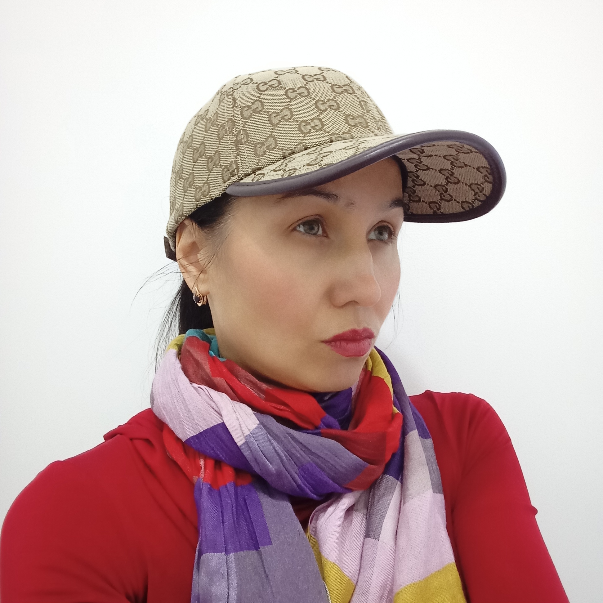 This is an eye-level, close-up portrait of a woman, possibly located in Izhevsk, Russia, set against a plain white background. The woman looks towards the right side of the frame with a thoughtful or pensive expression, her lips slightly pursed and colored with red lipstick. She has dark hair, partially visible, and wears a small gold-toned hoop earring with a dark accent in her visible left ear.
She is dressed in a vibrant red long-sleeved top and a colorful, checkered scarf wrapped around her neck, featuring blocks of red, purple, pink, yellow, and greyish-white. On her head, she wears a beige baseball cap with a repeating "GUCCI" monogram pattern, visible on both the top and the underside of the dark-rimmed visor. The lighting is bright and even, indicating an indoor setting.