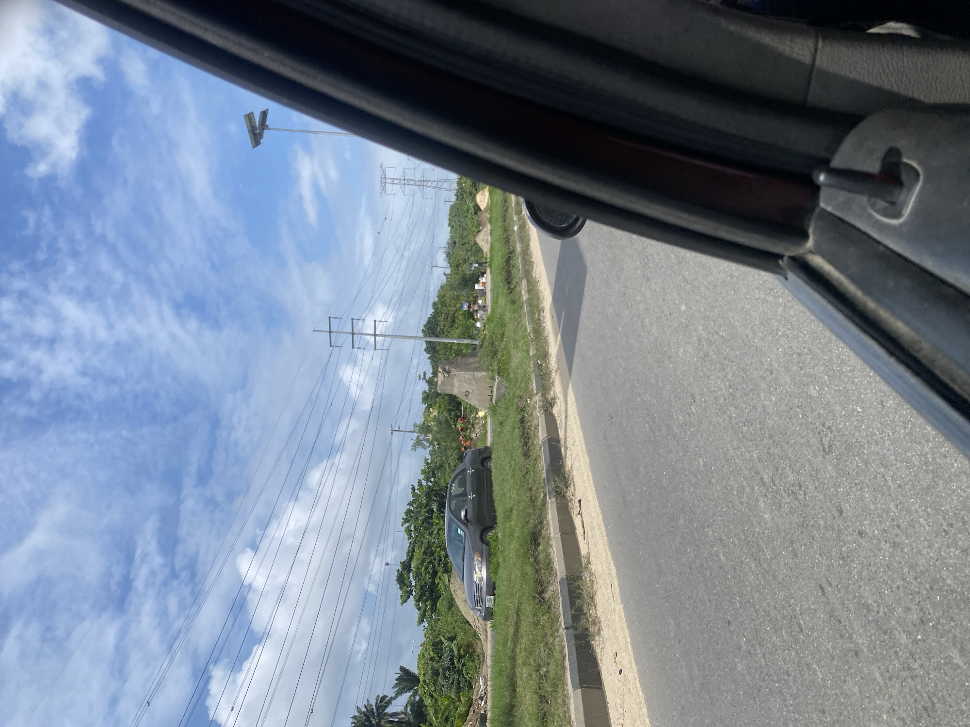 This is a medium-shot, eye-level, exterior shot taken from a vehicle, looking out towards the Lekki Peninsula II in Nigeria. The scene depicts a road with a gray asphalt surface, lined with a concrete curb and a grassy median. A dark gray sedan is parked on the median, facing left. Beyond the median, there are piles of sand, construction materials, and people in the distance, some of whom appear to be engaged in construction work. Overhead, numerous power lines stretch across the frame, supported by utility poles and pylons. The sky is predominantly blue with scattered white clouds, and palm trees are visible in the background. The right side of the frame is dominated by the interior of the vehicle, showing a portion of the car door and window frame.
