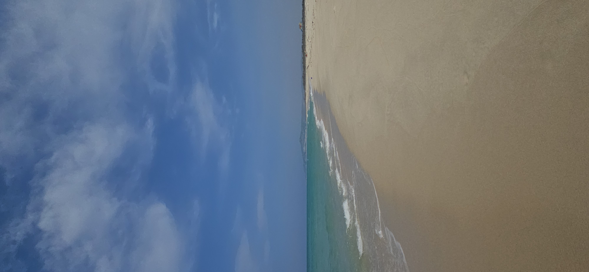 A tranquil landscape photograph depicts a sunny beach scene looking towards the ocean. The upper portion of the image features a bright blue sky with scattered white clouds, indicating clear weather. Below the sky, a vibrant turquoise ocean stretches, with gentle white waves breaking along the shore. A wide, light-brown sandy beach occupies the lower right section, showing subtle undulations and patterns from the water. In the distant background, a faint outline of hills or mountains is visible along the horizon line where the beach meets the sky. A few extremely small, indistinct figures or objects, possibly distant beachgoers or small colorful structures, can be barely perceived on the far end of the beach. The bright daylight suggests it is daytime, likely midday or early afternoon. This serene coastal view, set in Bac Cam Ranh Ward, Vietnam, evokes a sense of peace and natural beauty. There is no visible text.