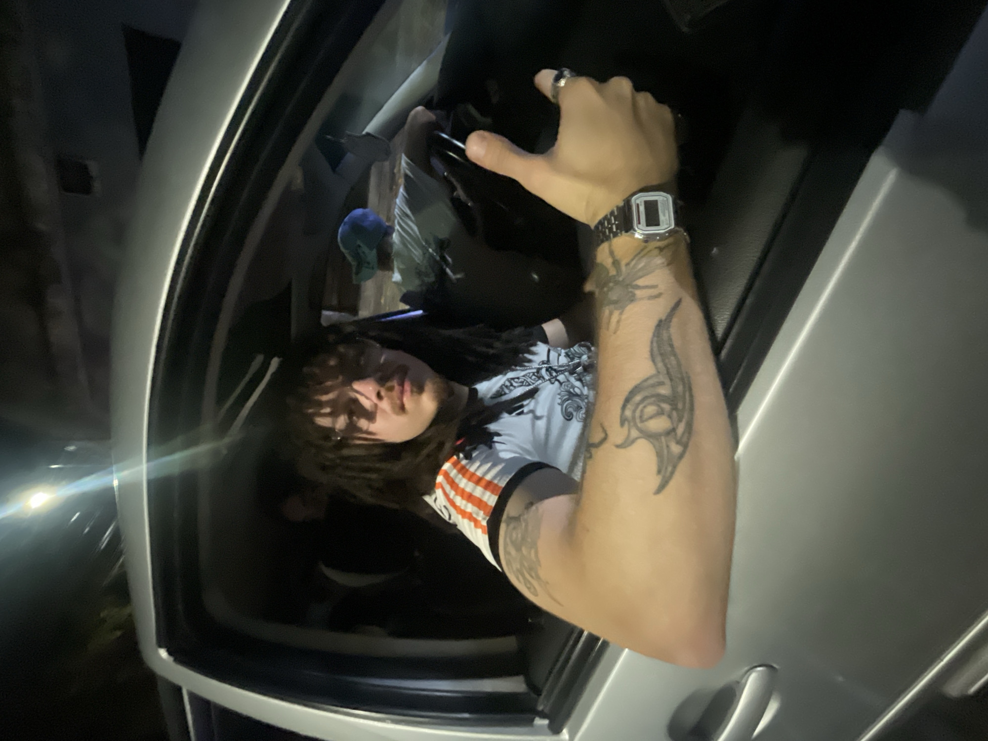 A person with dreadlocks is leaning out of a silver car, wearing a t-shirt with orange and white stripes on the sleeves. Their arm is extended, displaying a silver watch and a tattoo. The scene is dark, but a streetlight is visible in the background. The location is unknown and cannot be determined to be the city of Buenos Aires, Argentina from the image provided.