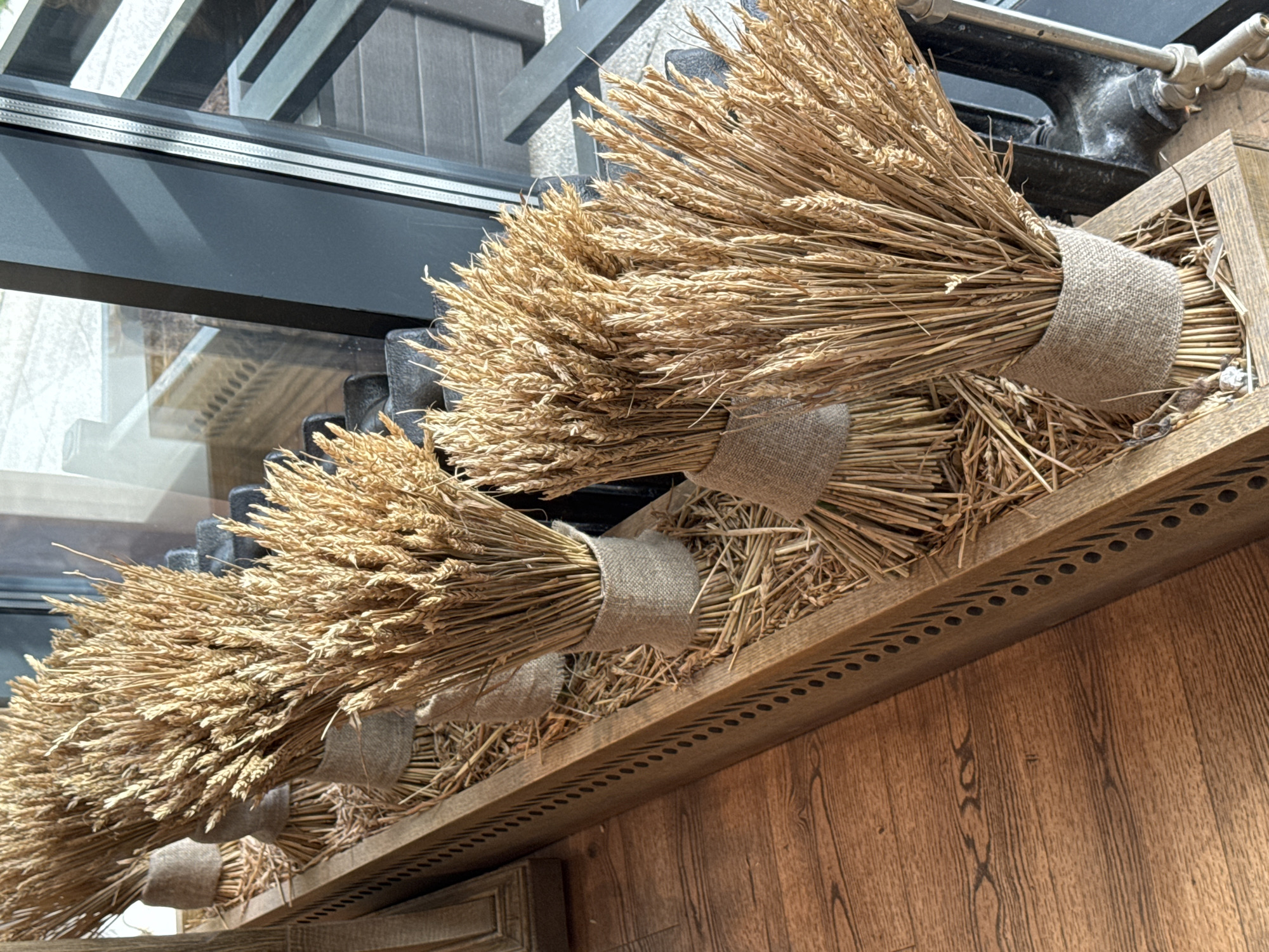 The image shows a decorative arrangement of wheat sheaves tied with burlap, displayed in a row along a wooden ledge. The sheaves are tightly bound, with the grain heads facing outward and upward. The wooden ledge is decorated with a pattern of alternating dots and dashes along its edge. A dark-colored window frame is visible in the background. The location is Izhevsk, Russia.