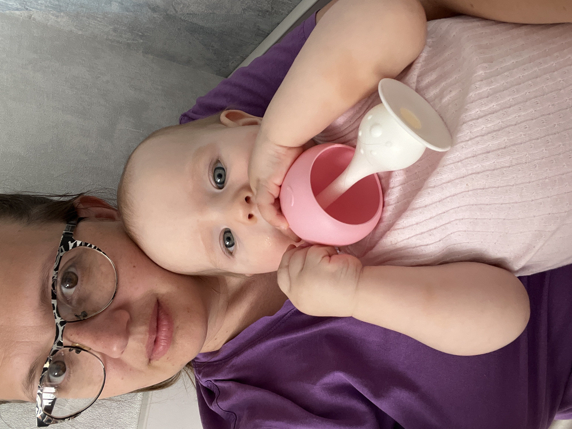 This media file features a woman and a baby lying down together, likely indoors in Chaikovsky, Russia. The image is taken from a top-down perspective, with both subjects oriented horizontally across the frame.
The woman, visible on the left side, has dark hair and is wearing distinct black and white patterned eyeglasses. Her eyes are a warm brown, and she has a neutral, calm expression. She is wearing a purple short-sleeved top.
Nestled beside her is a baby with light skin, blue eyes, and light hair, looking directly towards the viewer with a curious and engaged expression. The baby is wearing a light pink ribbed top. The baby is holding a bright pink sippy cup with a white, soft spout, bringing it to its mouth. Its hands are wrapped around the cup, suggesting it is either drinking or exploring the spout.
The background consists of a textured, light grey surface, possibly a wall or bedding. There are no specific cues for the time of day or weather. No visible text is present in the image. The scene conveys an intimate and calm moment between the mother and child.