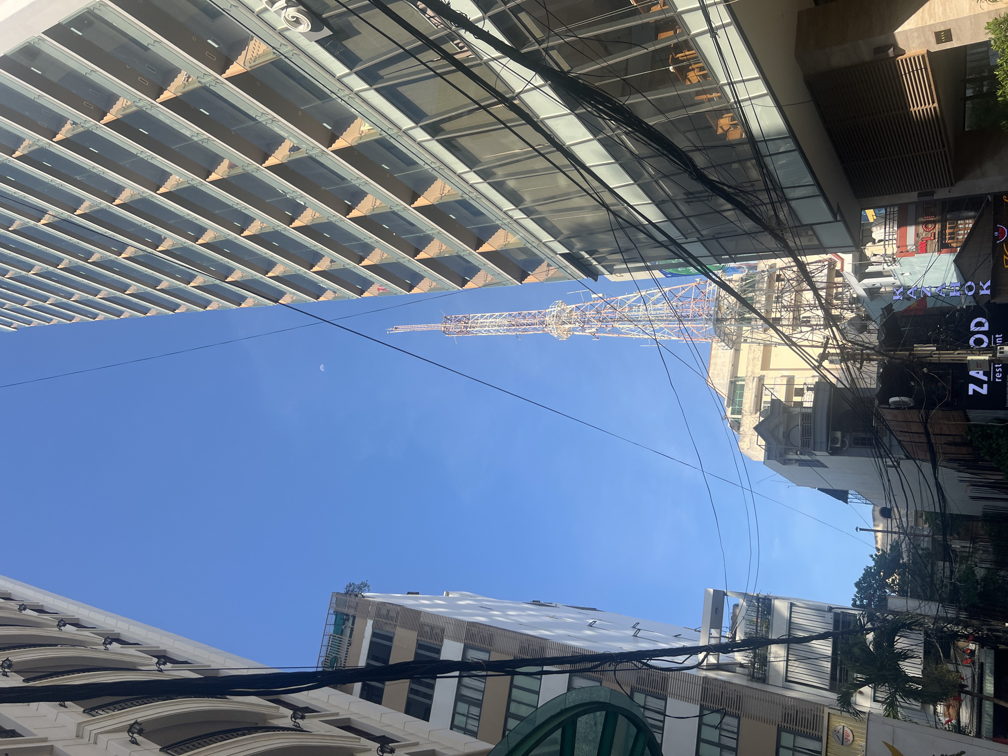 View looking up in the city of Nha Trang Ward, Vietnam. A tall metal tower stands in the middle of the frame. A blue sky is overhead with a small crescent moon visible. Buildings rise on either side of the tower. On the left, a building with a modern glass facade is visible. On the right, a building with shops and apartments is visible, along with utility wires. At the bottom of the frame, the tops of buildings are visible.