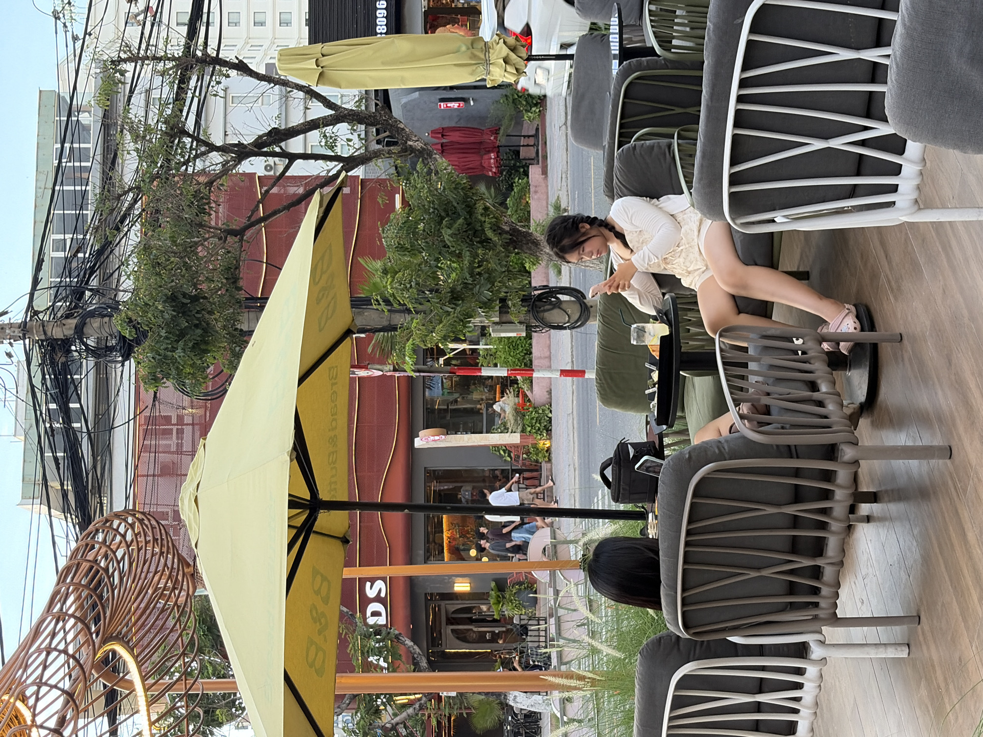 The image depicts an outdoor seating area of a cafe in Da Nang, Vietnam, likely on a street with commercial buildings and overhead electrical wires. In the foreground, two women are seated on chairs. The woman on the right, with her back to the camera, is wearing a white top and shorts, holding a pink smartphone and looking down at it. Her legs are crossed, and she is wearing pink clogs. The woman on the left, also with her back to the camera, has dark hair and is wearing a black top. She is seated at a table with a laptop and a drink.
Behind them, a large yellow umbrella provides shade. The cafe's signage, partially obscured by the umbrella, appears to say "Lands" in white letters on a dark background, with red accents on the building's facade. Several people are walking on the sidewalk in front of the cafe. Trees and potted plants are visible, adding greenery to the urban setting. A street is visible behind the sidewalk, with a white car parked further down. The sky is overcast. The overall scene suggests a casual daytime or early evening setting in a bustling city.