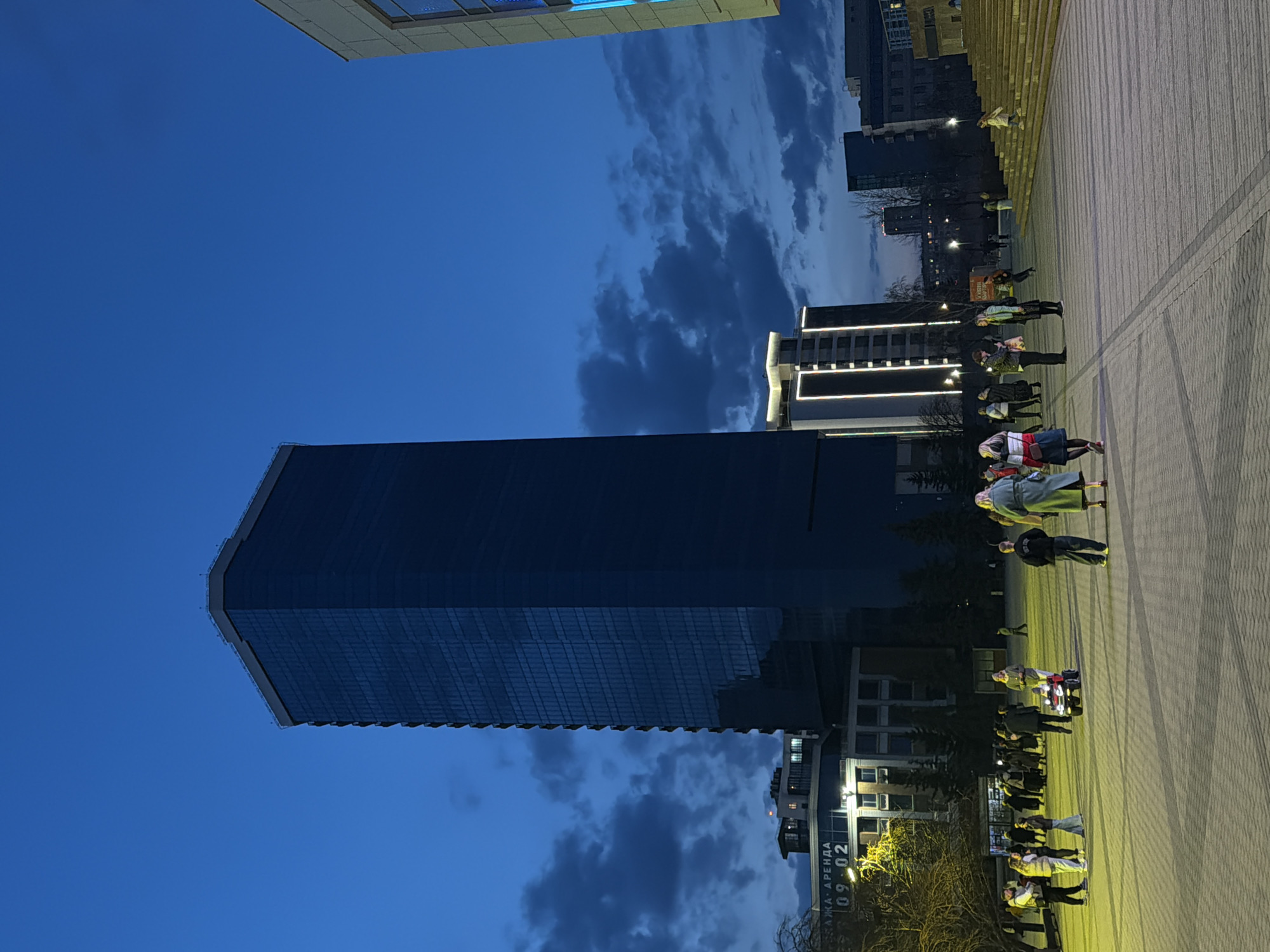 The image presents an urban cityscape at dusk or early evening in Krasnoyarsk, Russia. A tall, dark, modern skyscraper with a reflective glass facade dominates the left side of the frame, standing against a deep blue sky. The sky above features scattered dark clouds, indicative of the fading daylight. To the right and in the background, several other city buildings are visible, some illuminated with internal lights or decorative facade lighting.
The foreground and right portion of the image consist of a wide, paved pedestrian area or public square, likely made of interlocking bricks or patterned concrete. Numerous individuals are scattered across this area, some walking and others standing in small groups. They are illuminated by artificial light sources, casting visible light on the ground and figures. On the far right, a set of broad steps or a tiered structure rises. Bare trees are visible between the buildings in the mid-ground.
On a sign or light pole located in the middle-right section of the pedestrian area, Cyrillic text is discernable. The words "АРЕНДА" (meaning "Rent") are clearly visible, along with the numbers "09.02". The overall scene depicts a modern city environment during its evening transition, with active public spaces and prominent architecture.